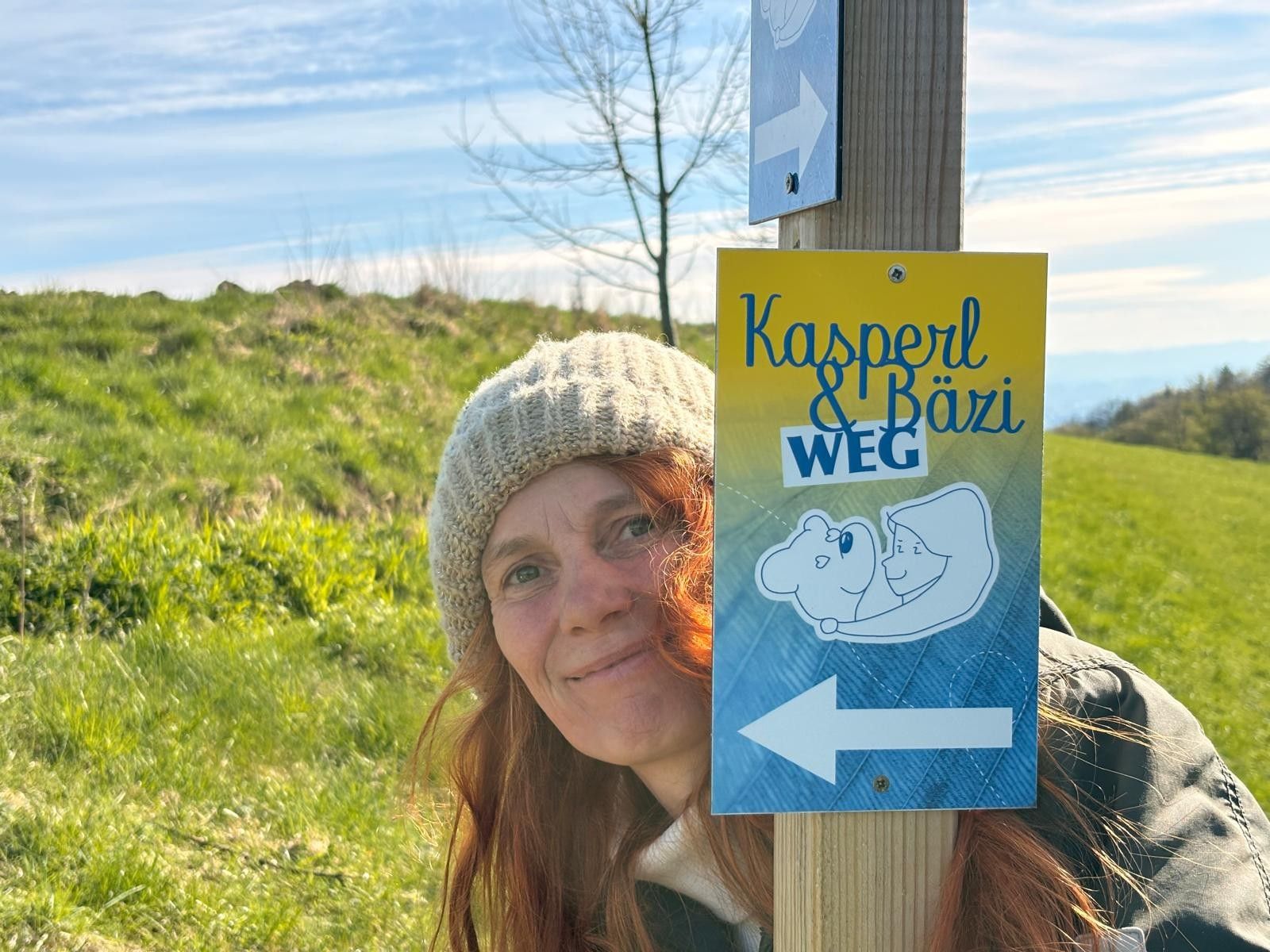 Egy sapkás nő áll egy „Kasperl & Bäzi Weg” feliratú útjelzőtábla mellett egy zöld tájban.