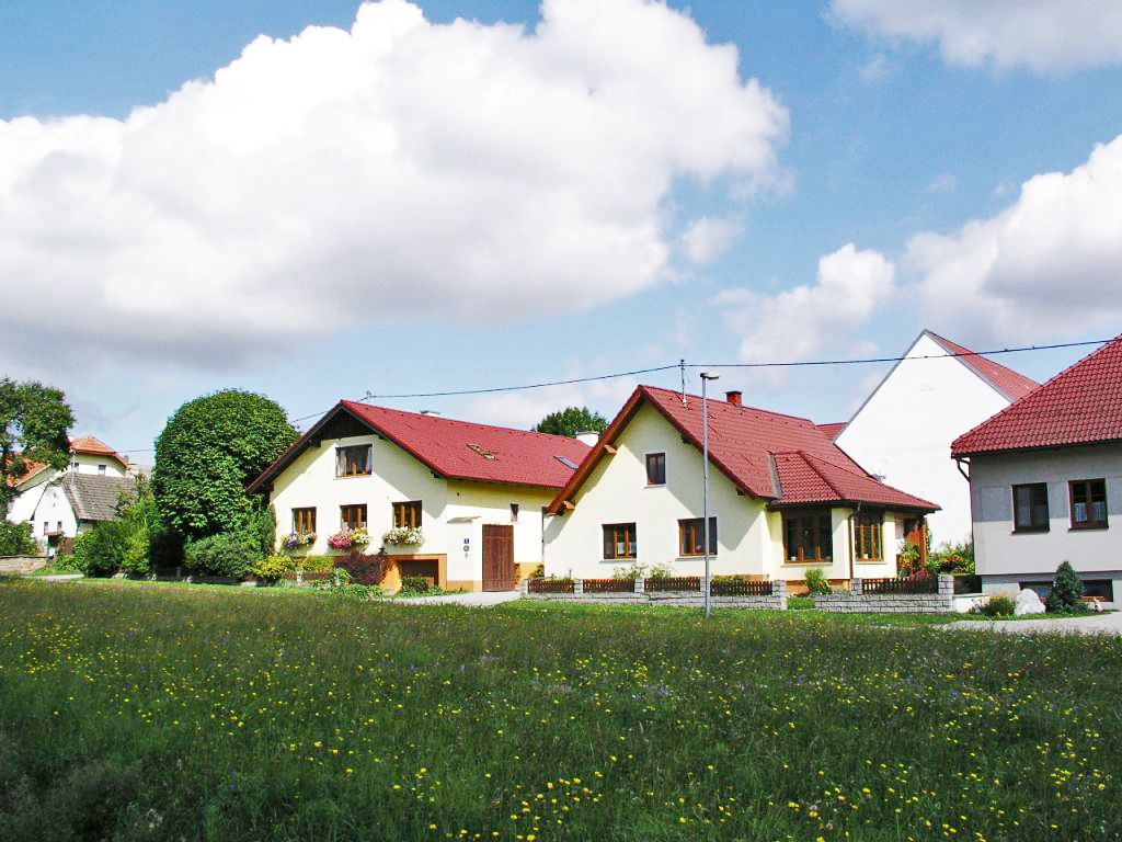 Egy biofarm, több sárga épülettel és piros tetővel, zöld rétekkel és fákkal körülvéve, kék felhős égbolt alatt.