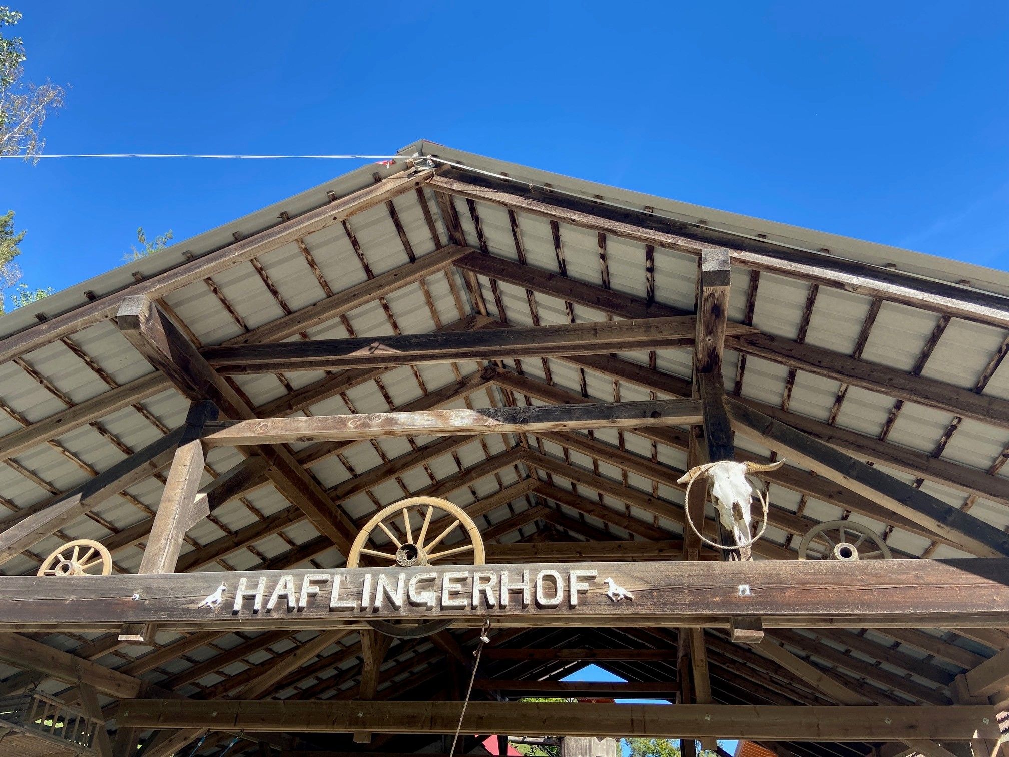 Egy faépület teteje "Haflingerhof" felirattal, valamint szekérkerekeket és állatfejet ábrázoló díszítésekkel.