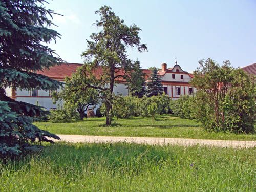 Egy történelmi épület piros tetővel, fákkal és zöld réttel körülvéve.
