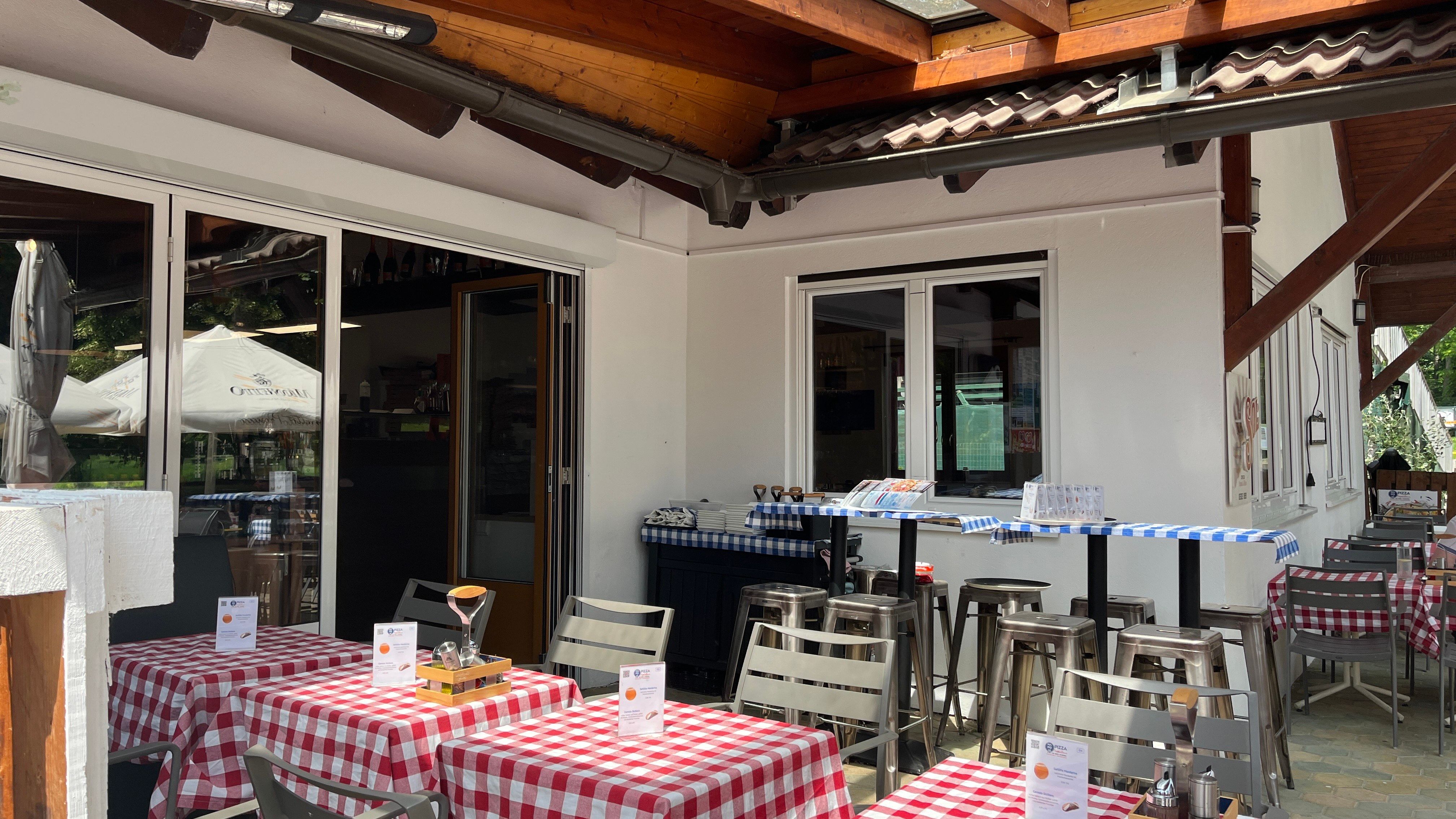 A pizzéria bejárata a vendégkerttel. Az előtérben piros terítővel letakart asztalok.