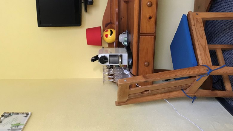 Egy kis faasztal, k&aacute;v&eacute;főző, poharak, egy l&aacute;mpa &eacute;s egy telev&iacute;zi&oacute; a falon.