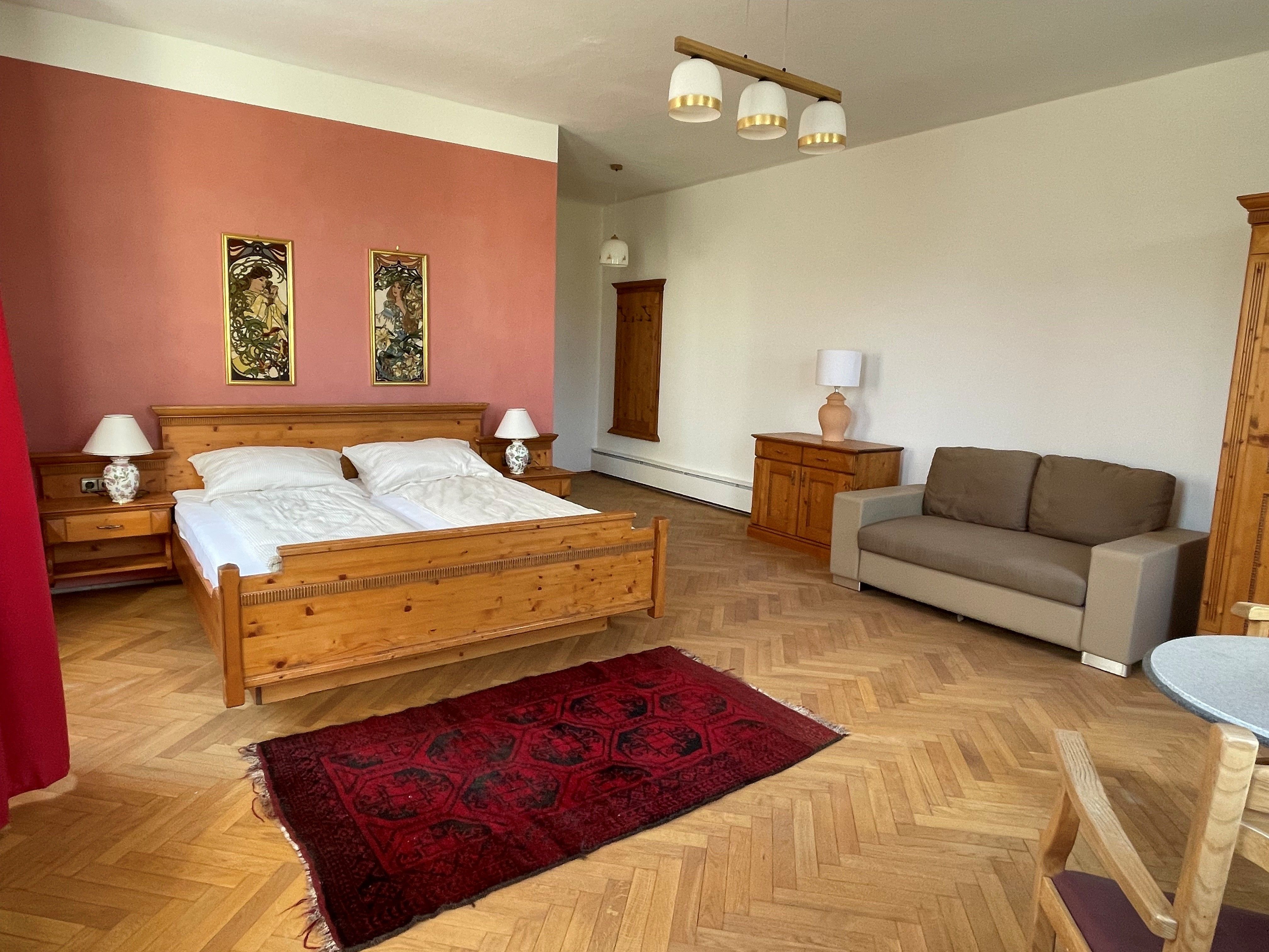 Egy tágas hálószoba, amelyben egy faágy, éjjeliszekrények, egy kanapé és egy piros szőnyeg található a fa padlón.