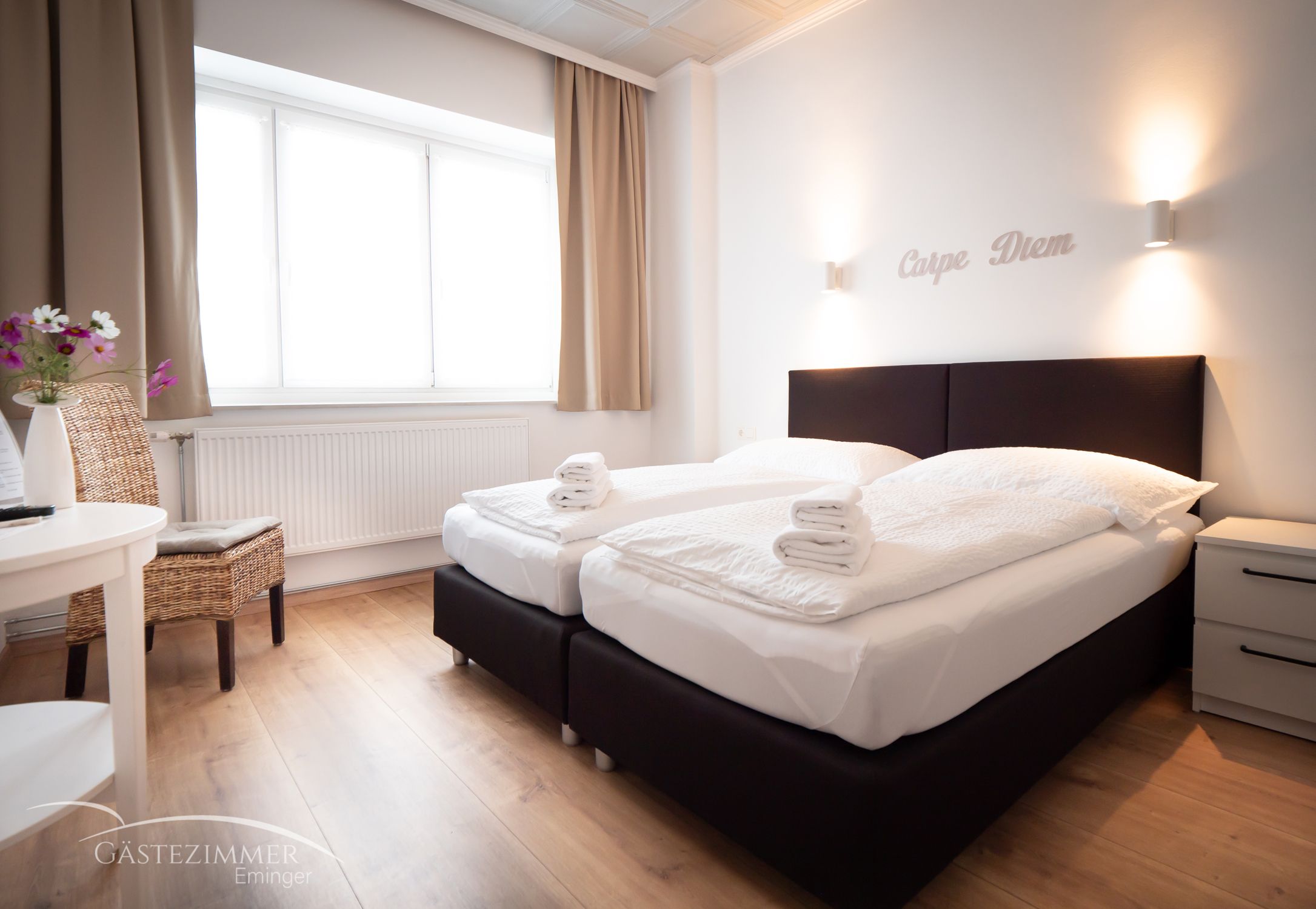 Modern kétágyas szoba két ággyal, fehér paplanokkal és törölközőkkel. "Carpe Diem" felirat a falon, világos függönyök és fapadló.