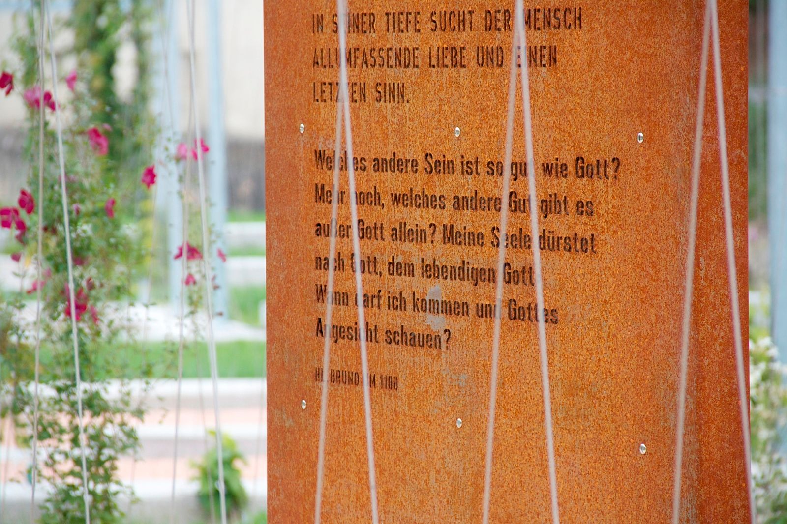 Közelkép egy rozsdás fémtábláról, vésett szöveggel, növényekkel körülvéve.