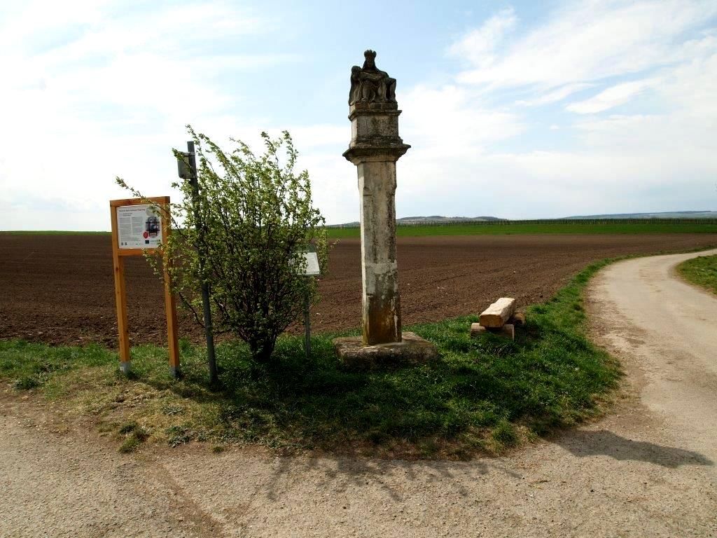 Kőoszlop egy országút mellett, információs táblával és bokrokkal.