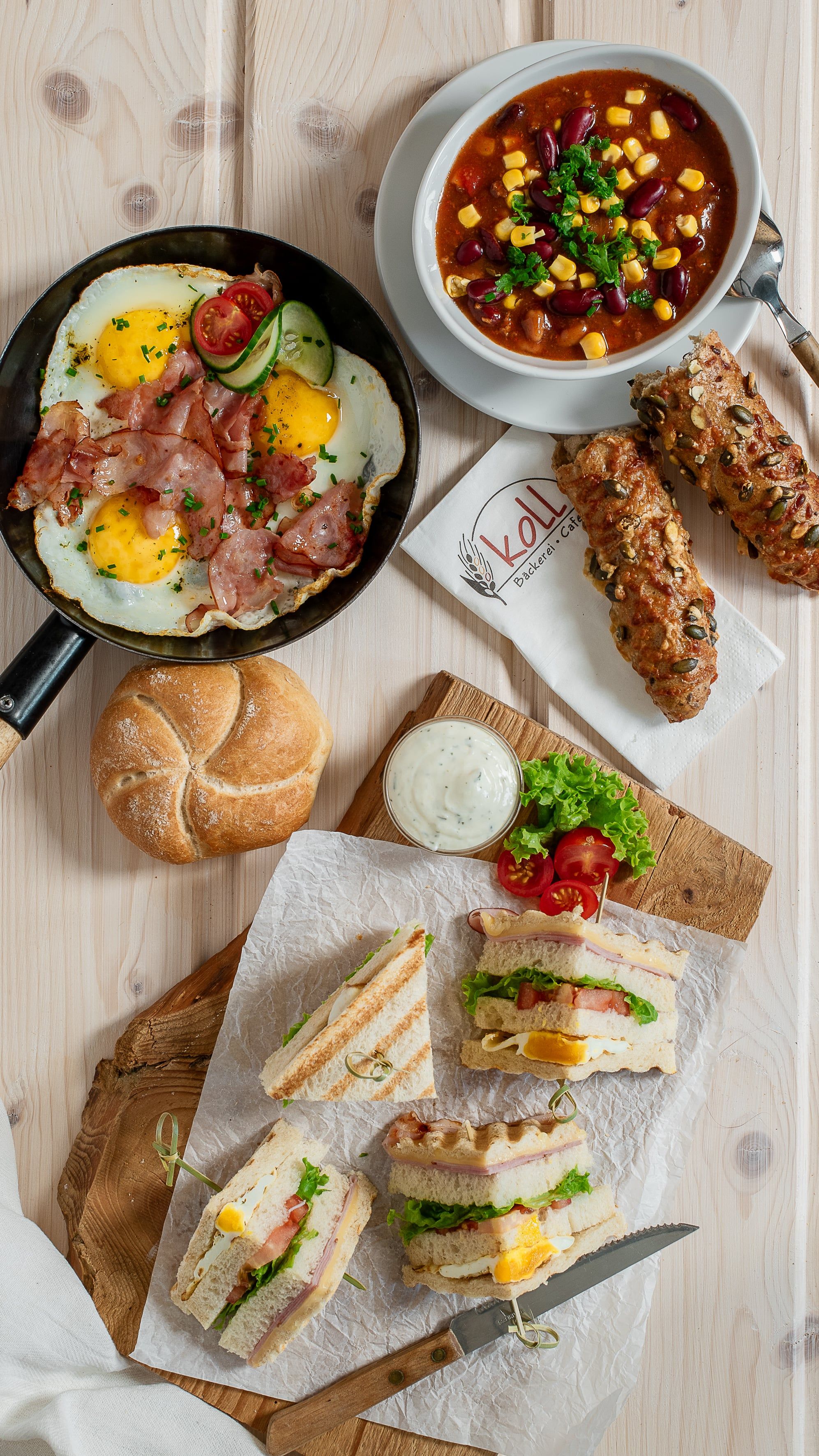 Különböző meleg harapnivalók egy asztalon, többek között tükörtojás szalonnával, leves, szendvicsek és zsemle.