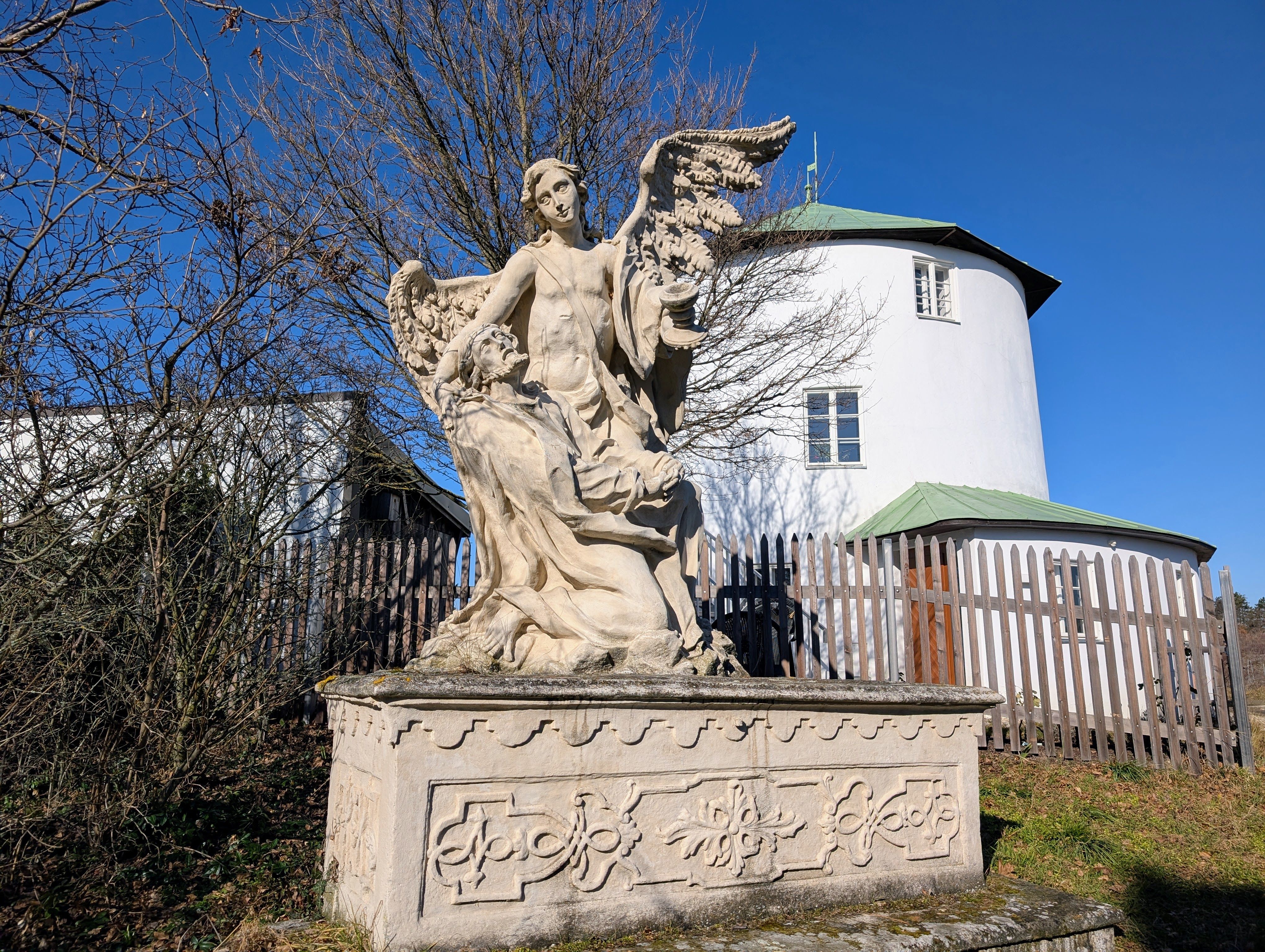 Kőszobor, szárnyas angyal és térdelő alak egy kerek, fehér, zöldtetős épület előtt.