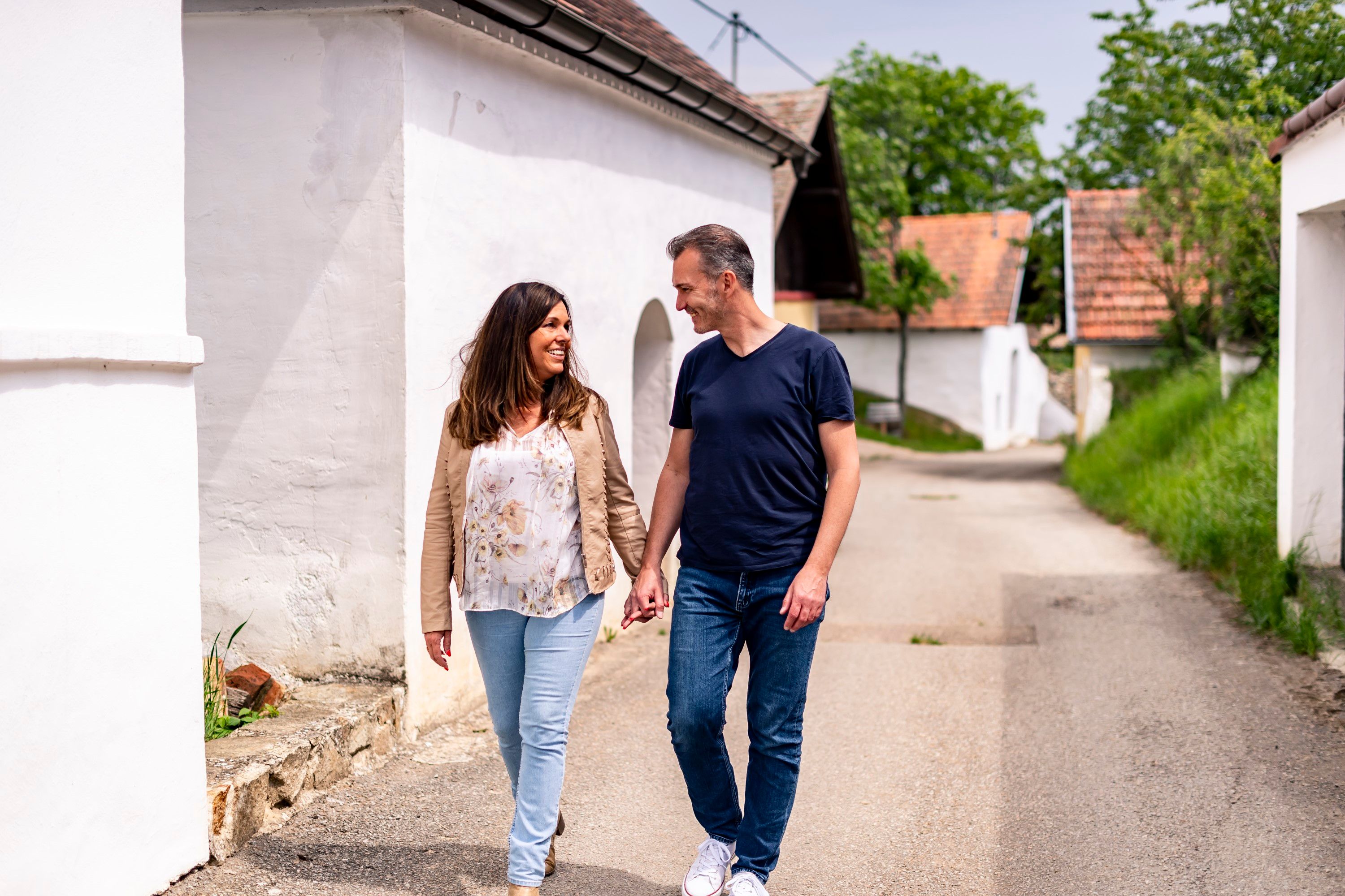 Egy pár kéz a kézben sétál a pillersdorfi Öhlbergkellergasse-on, amelyet hagyományos borospincék és zöld vidék vesz körül.