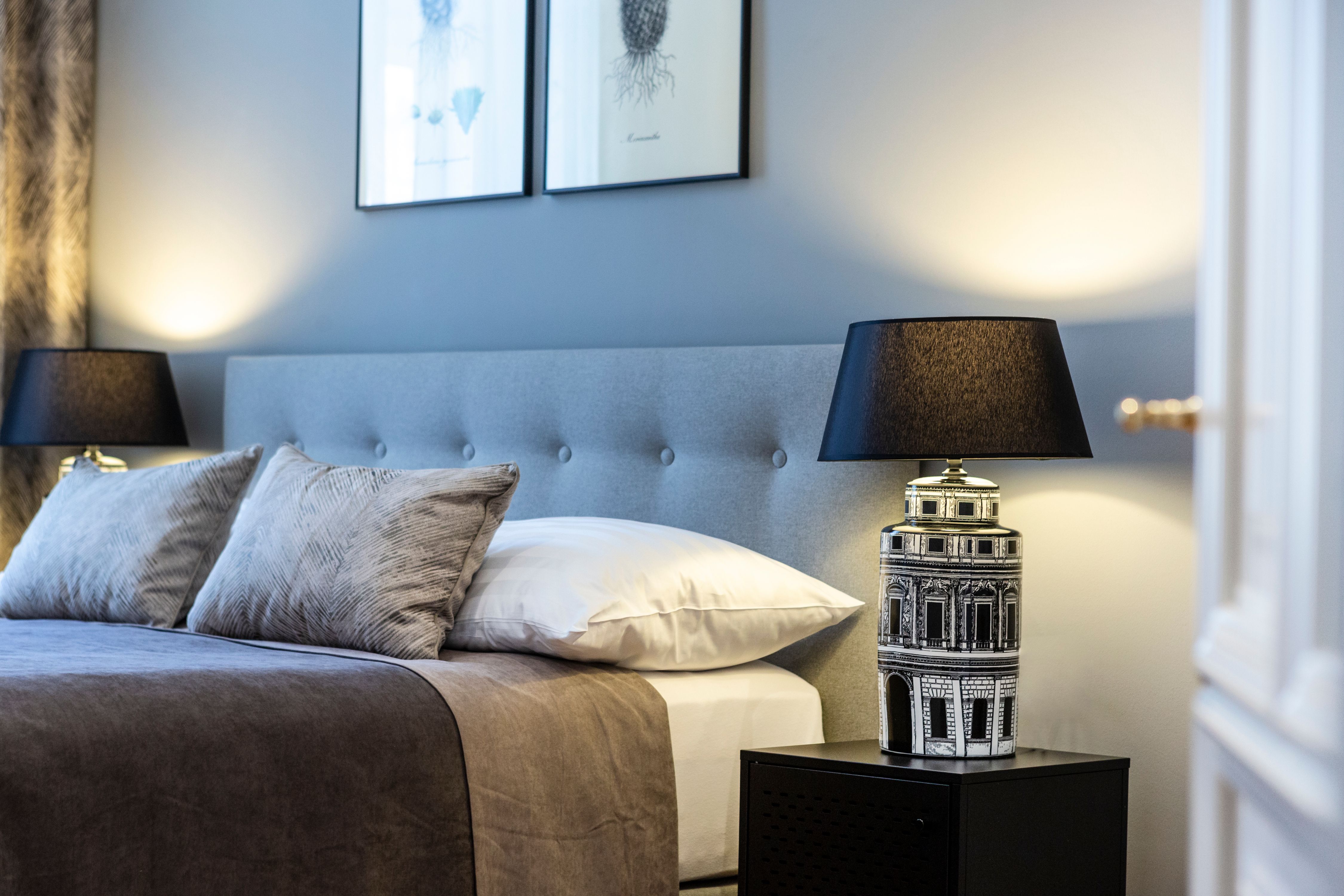 Elegáns kétágyas szoba szürke fejvéggel, két párnával, éjjeli lámpával és faliképekkel.