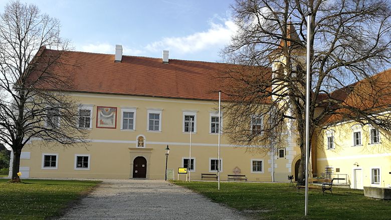 Sárga kastélyépület piros tetővel és toronnyal, fákkal körülvéve.