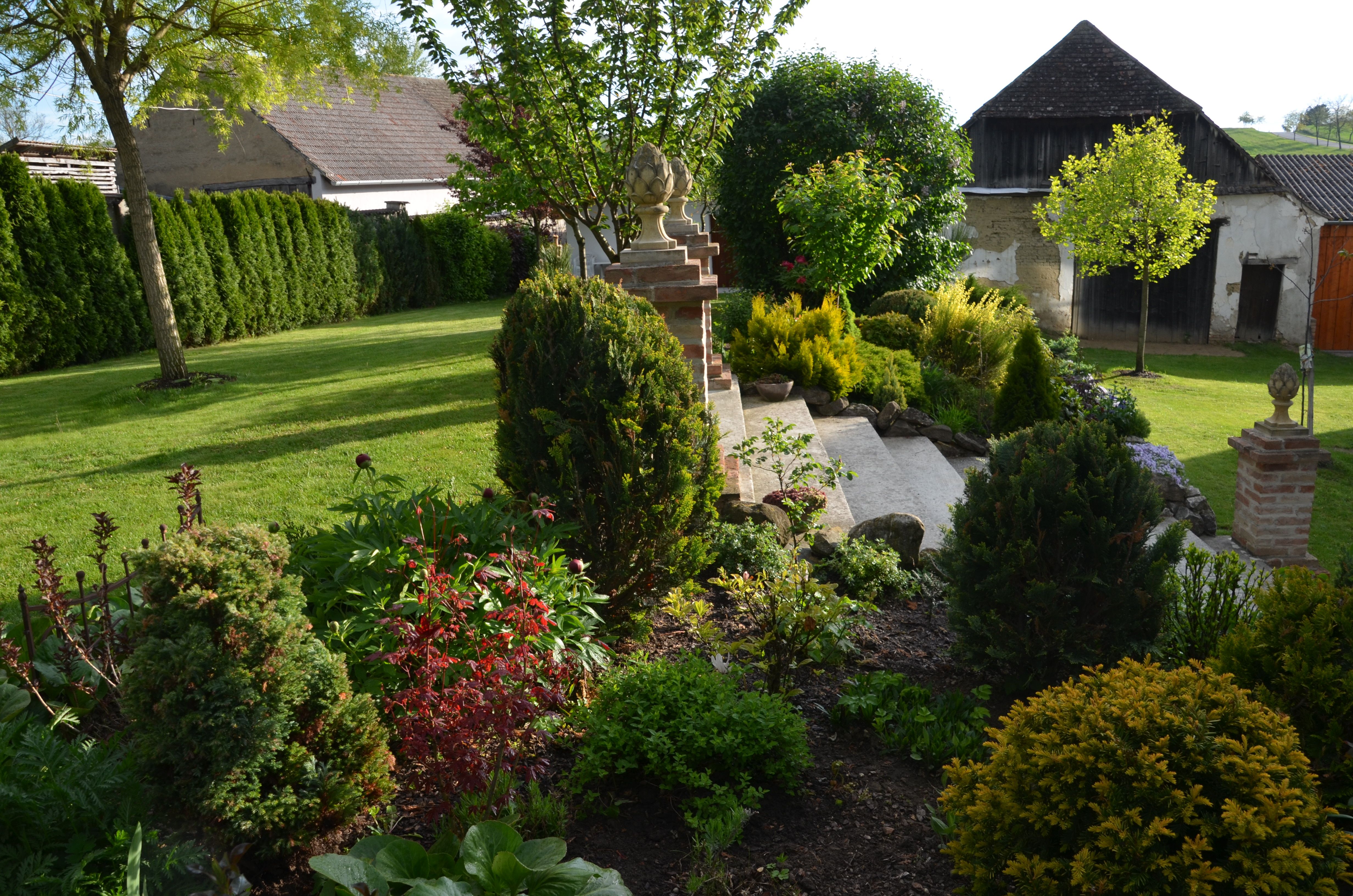 Gondozott kert gyeppel, bokrokkal és fákkal, a háttérben épületekkel körülvéve.