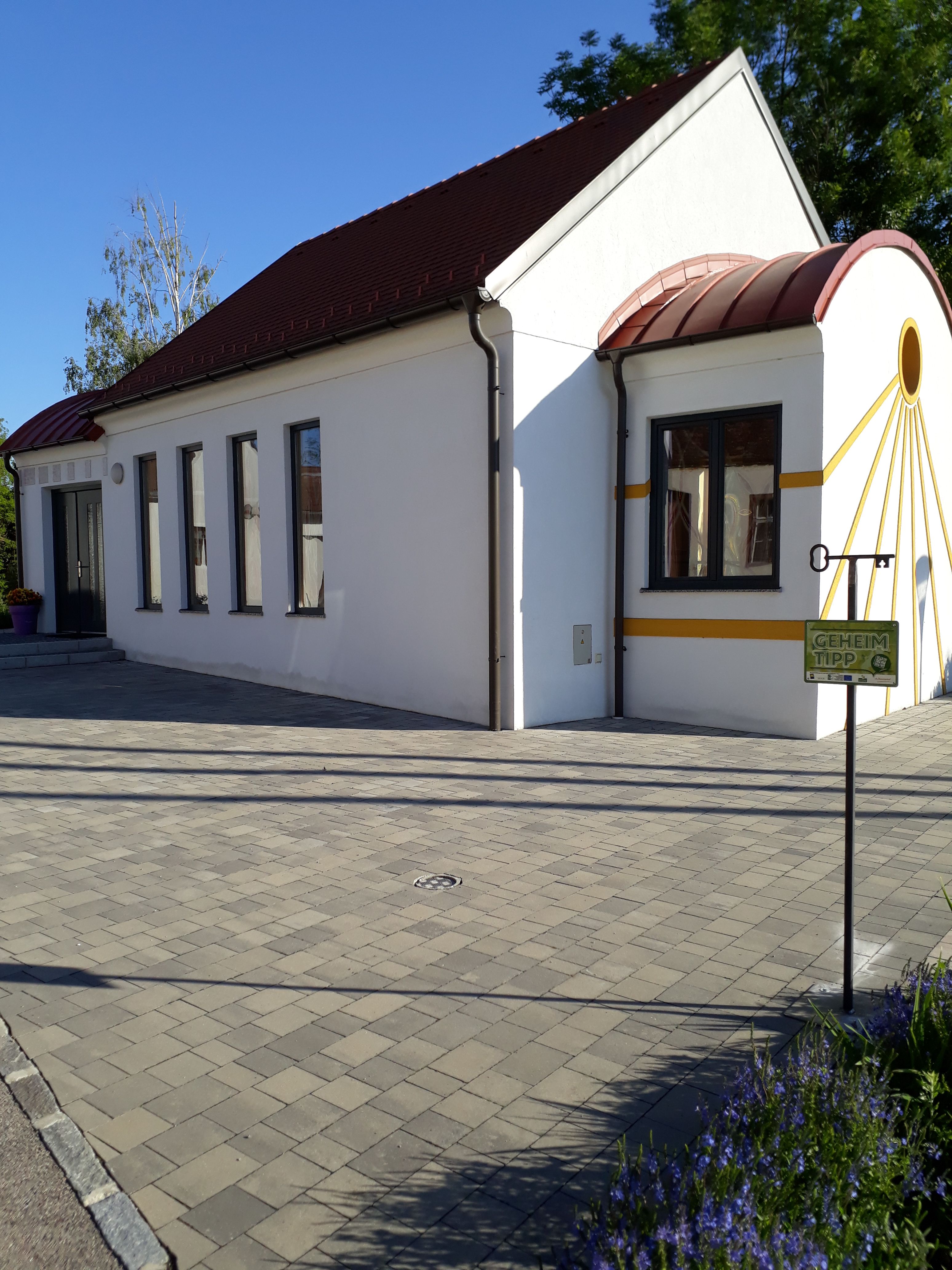 Fehér kápolna piros tetővel és sárga akcentussal, körülötte térkővel burkolt padló és növények.