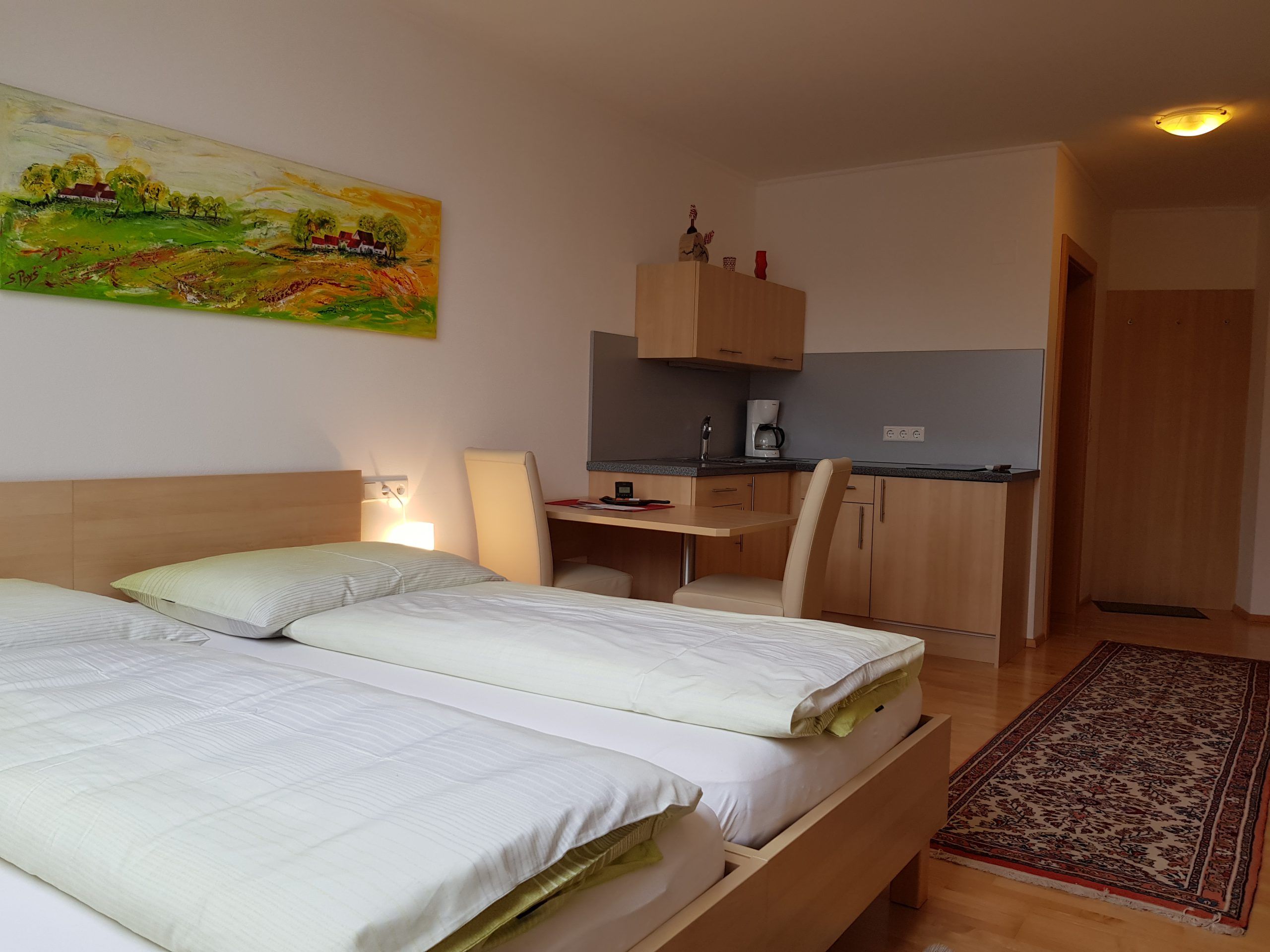 Modern szoba két ággyal, íróasztallal, kis konyhasarokkal és egy színes festménnyel a falon.