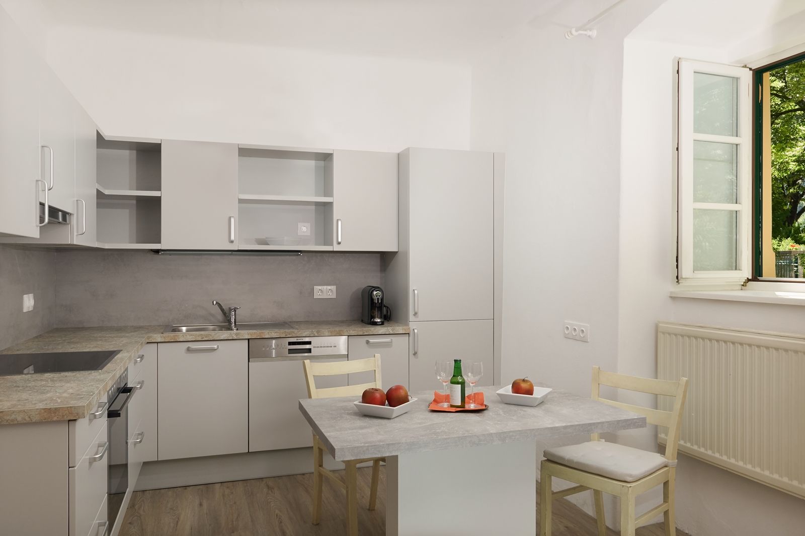 Modern konyha szürke szekrényekkel, asztal almával és itallal, nyitott ablak.