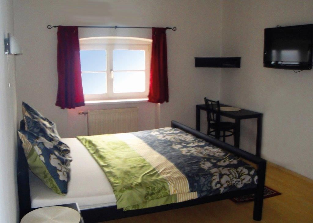 Egy hálószoba egy ággyal, egy vörös függönyös ablakkal, egy kis asztallal és székkel, valamint egy falra szerelt televízióval.