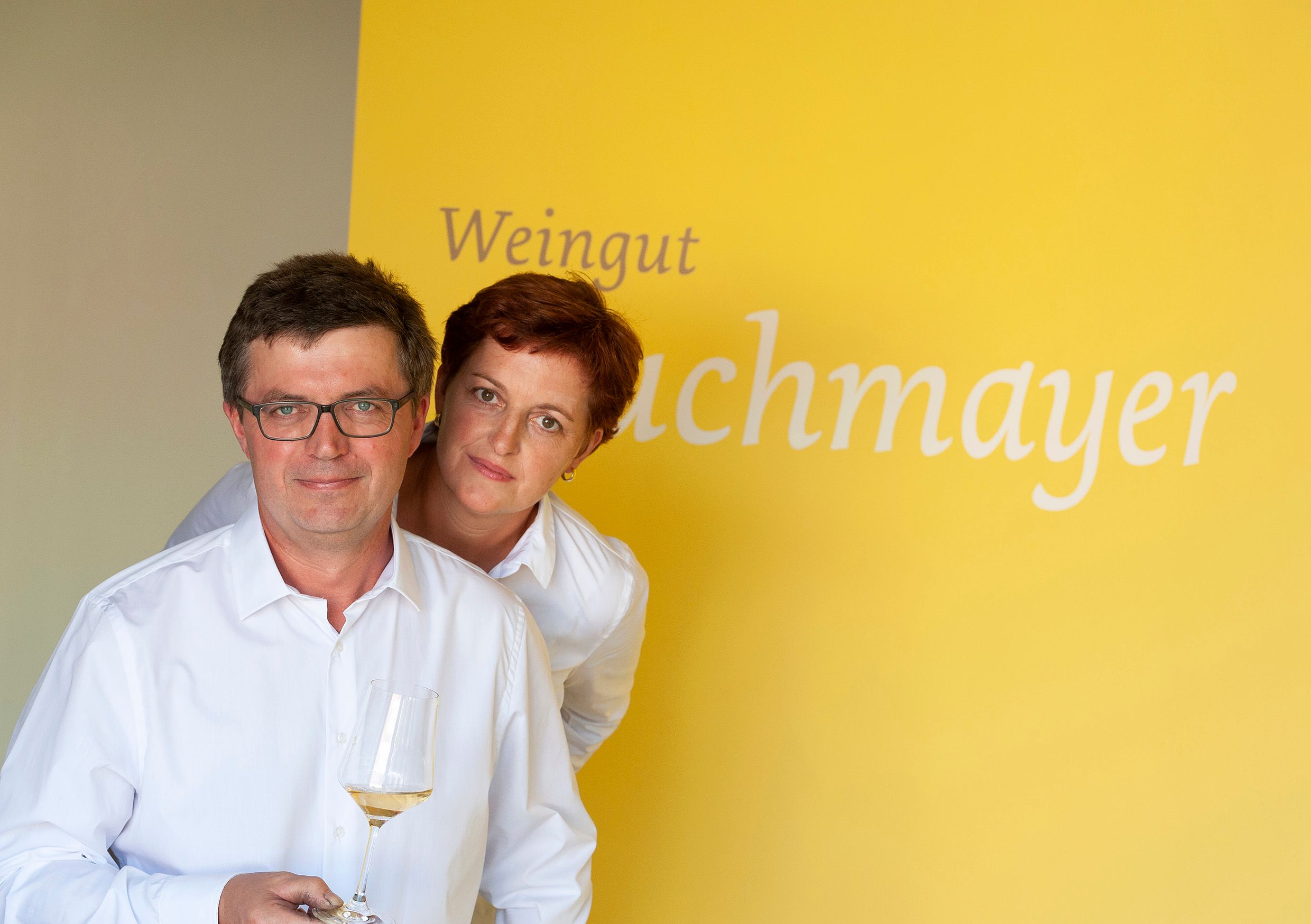 Két fehér inges ember sárga háttér előtt, "Weingut Buchmayer" felirattal. Az egyik személy egy borospoharat tart a kezében.