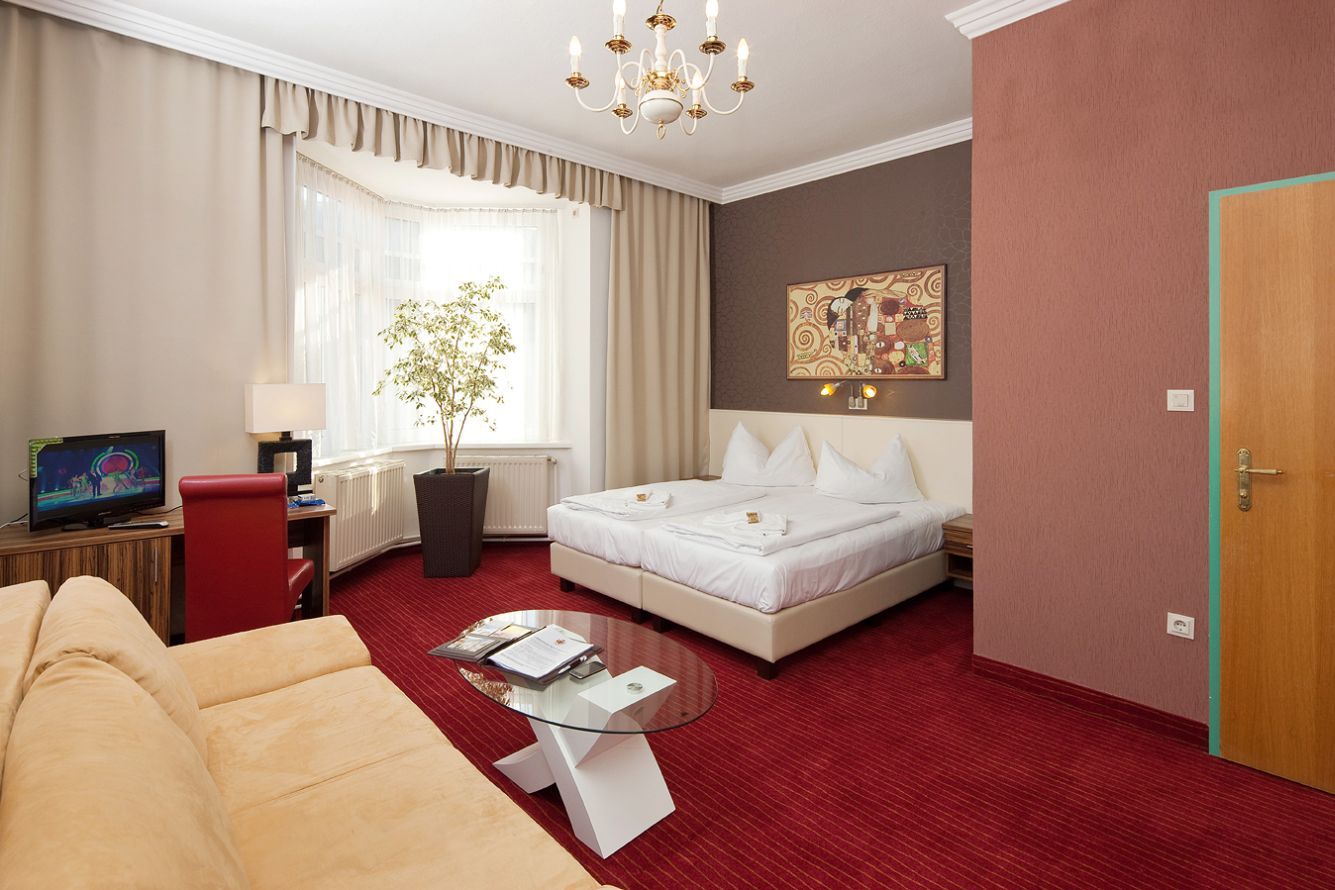 Hotelszoba franciaággyal, vörös szőnyeggel, kanapéval, TV-vel és nagy ablakkal.