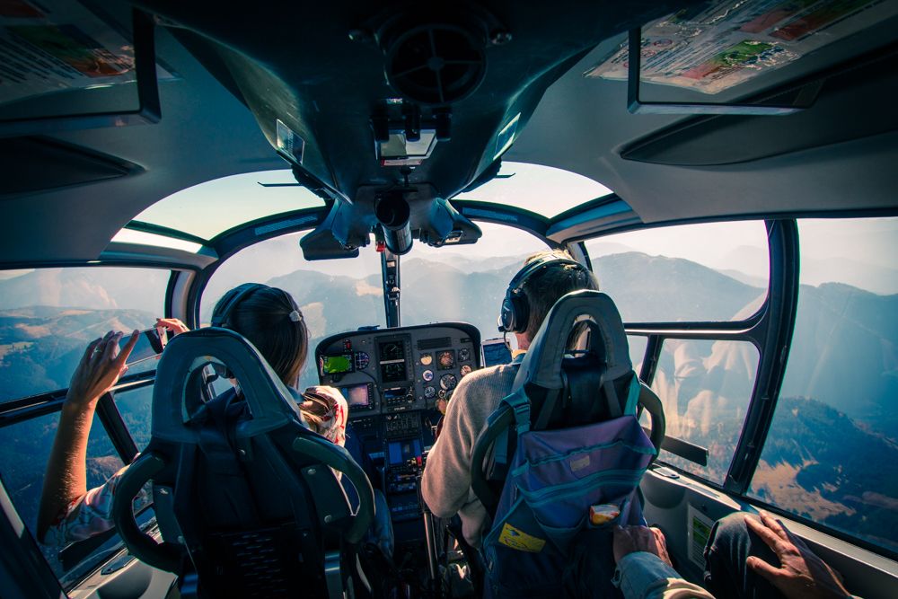 Kilátás egy helikopter pilótafülkéjéből, két ember repül egy hegyvidéki táj felett.