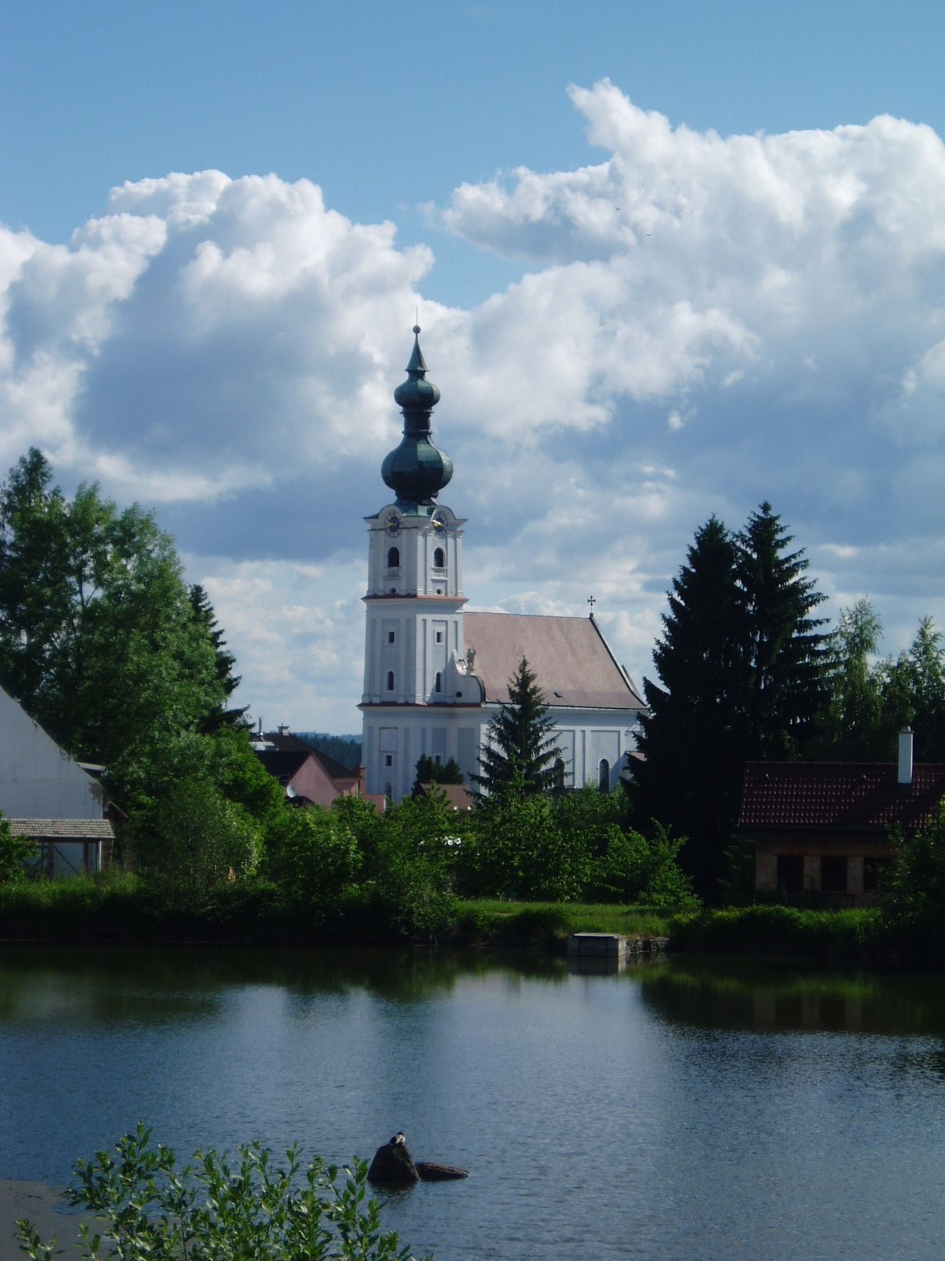Templom Kirchberg am Walde-ban, előtérben egy tó, felhős égbolt.