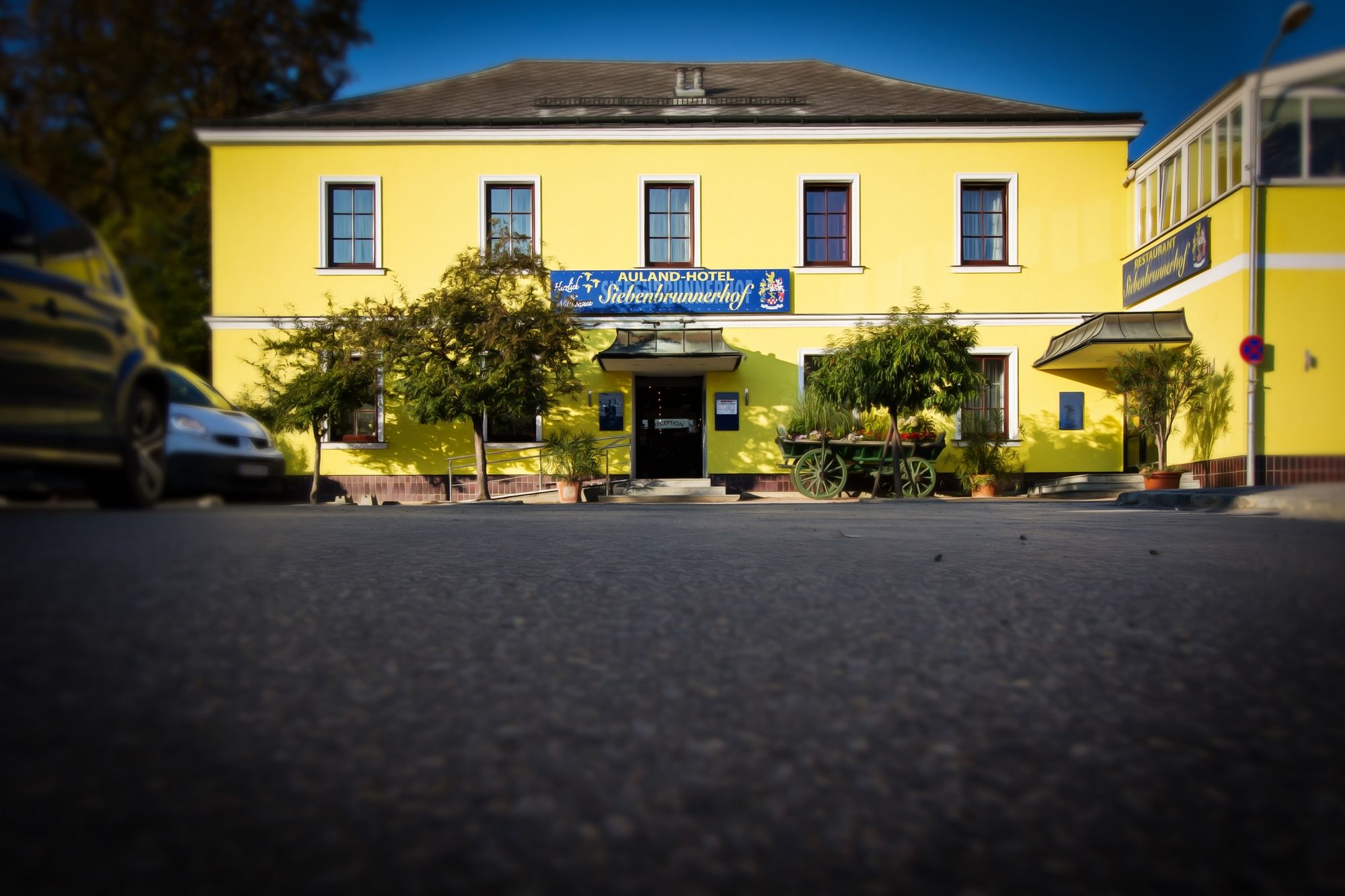 "Auland-Hotel Siebenbrunnerhof" feliratú sárga épület, fákkal és egy régi kocsival körülvéve.