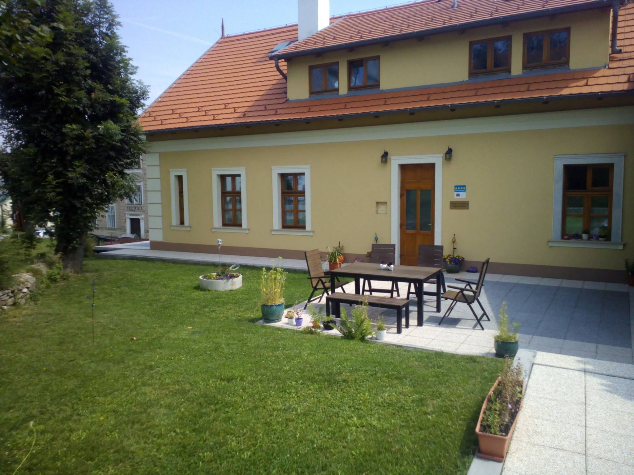 Sárga ház piros tetővel és terasszal, pázsittal és növényekkel körülvéve.