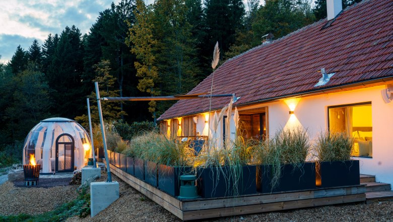 Csodákkal teli faházak és helyek - Weidehaus De Luxe, © WUNDERVOLL eben Egy modern faház terasszal és egy átlátszó kupolás sátorral az erdőben alkonyatkor.