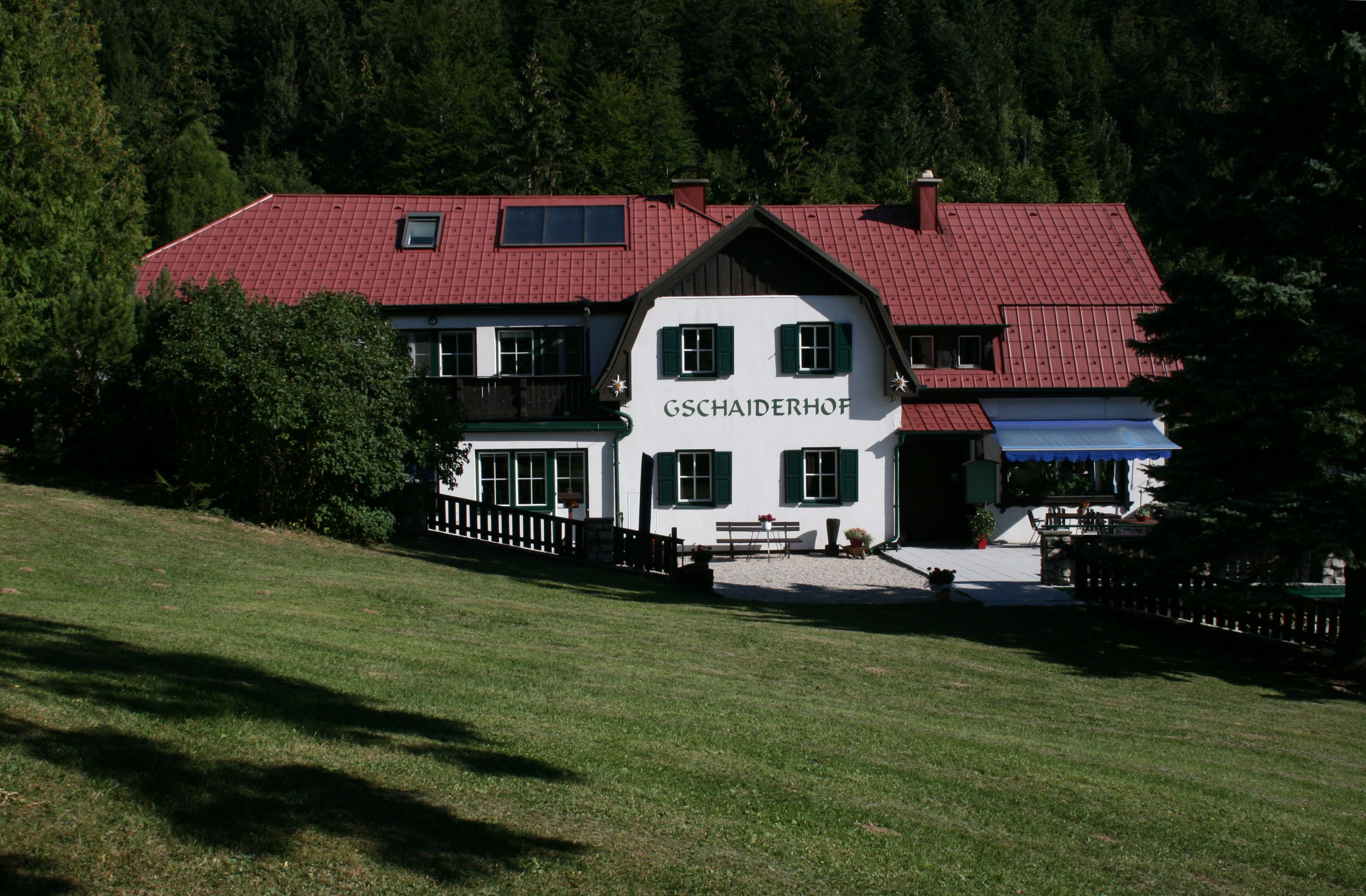 Hagyományos épület piros tetővel és zöld redőnyökkel, fákkal és rétekkel körülvéve, a "Gschaiderhof" felirattal.