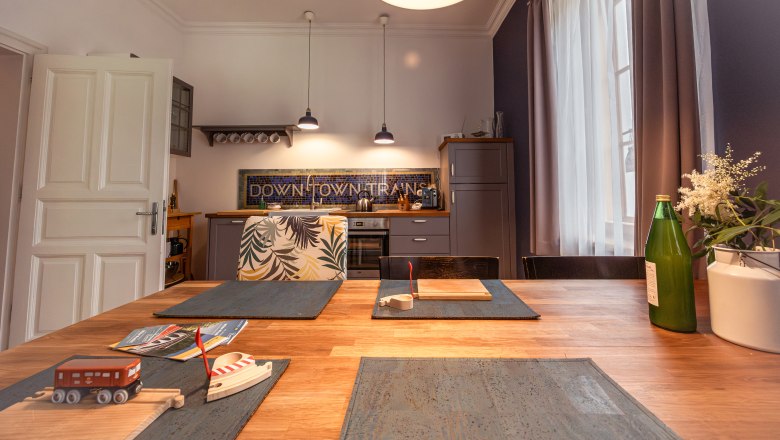 Konyha fa asztallal, j&aacute;t&eacute;k vonattal, &uuml;veggel &eacute;s vir&aacute;gokkal az asztalon, a h&aacute;tt&eacute;rben modern l&aacute;mp&aacute;k &eacute;s konyhaszekr&eacute;nyek.