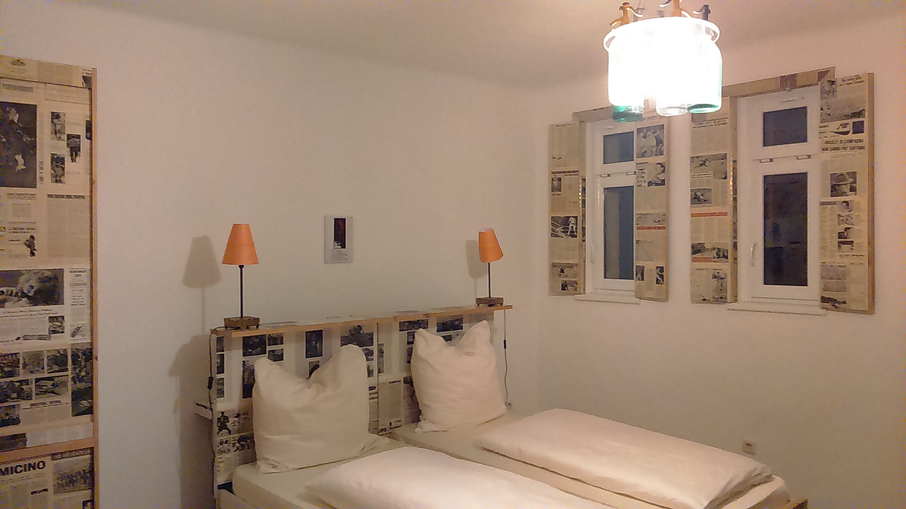 Hotelszoba franciaággyal, a falakon és az ablakokon újságdíszítés, két narancssárga színű lámpa.