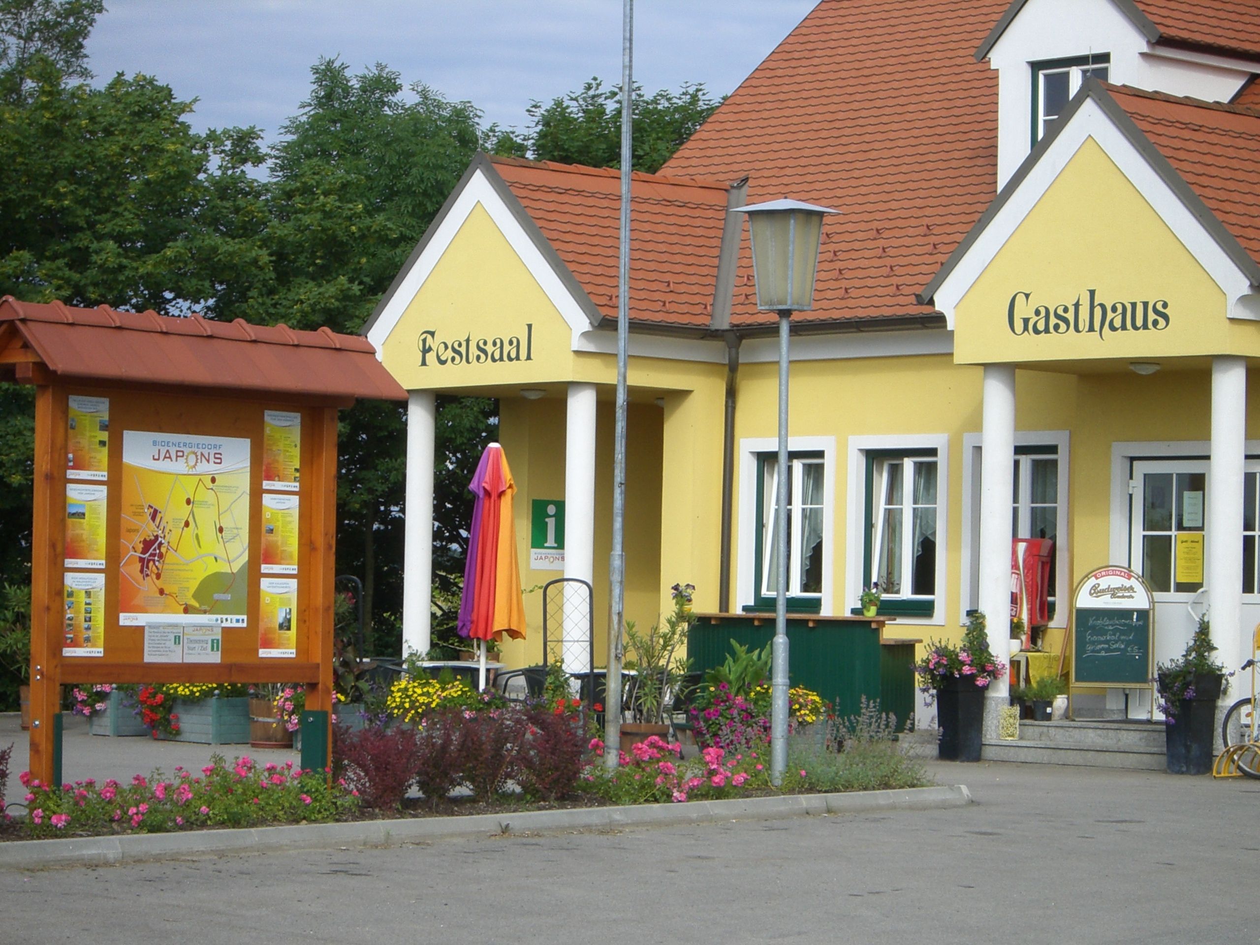 Sárga színű, "Festsaal" és "Gasthaus" feliratú épület, virágokkal és információs táblával körülvéve.