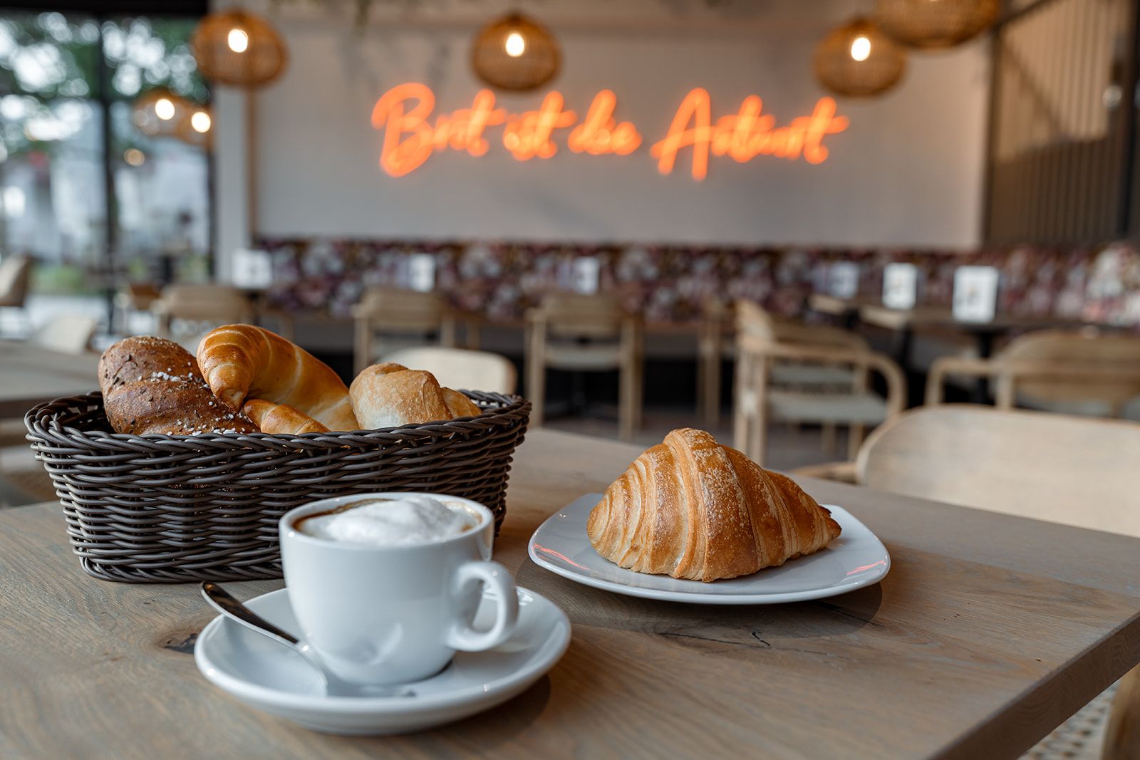 Egy kávézó, egy kosár sütemény, egy croissant egy tányéron és egy csésze cappuccino egy asztalon.