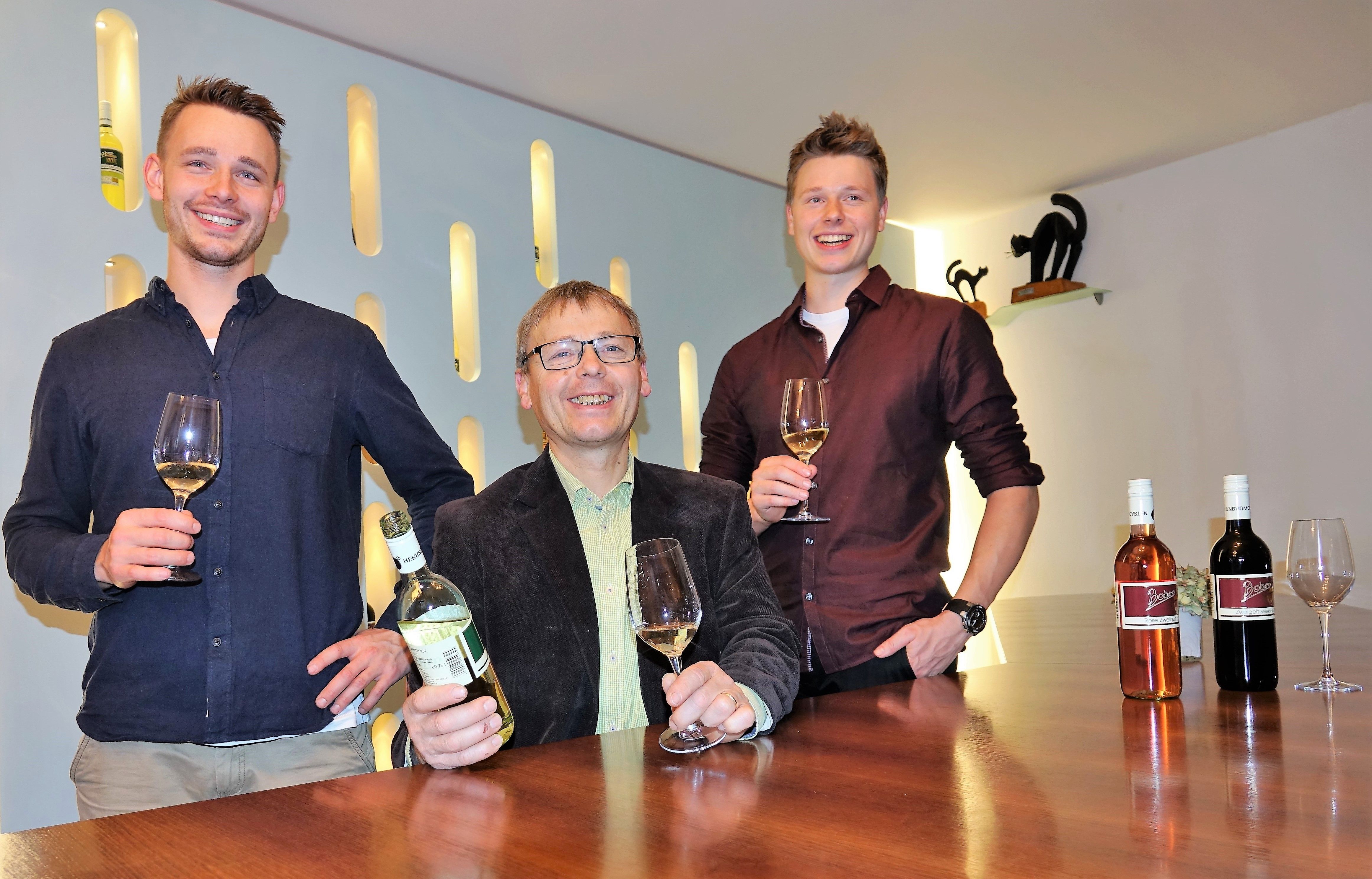 Három férfi áll egy asztalnál borospoharakkal és borosüvegekkel.
