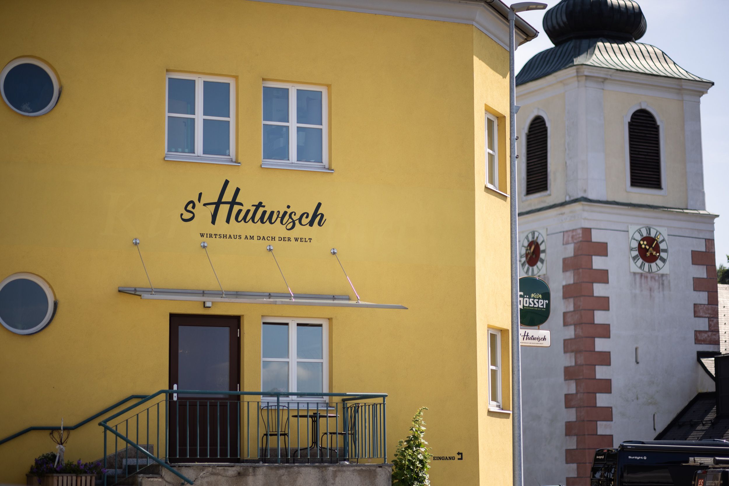 Sárga színű, "s'Hutwisch" feliratú épület egy órával ellátott templomtorony mellett.