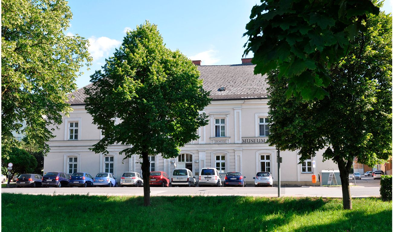 Egy fehér, "Múzeum" feliratú épület, körülötte fák és parkoló autók.