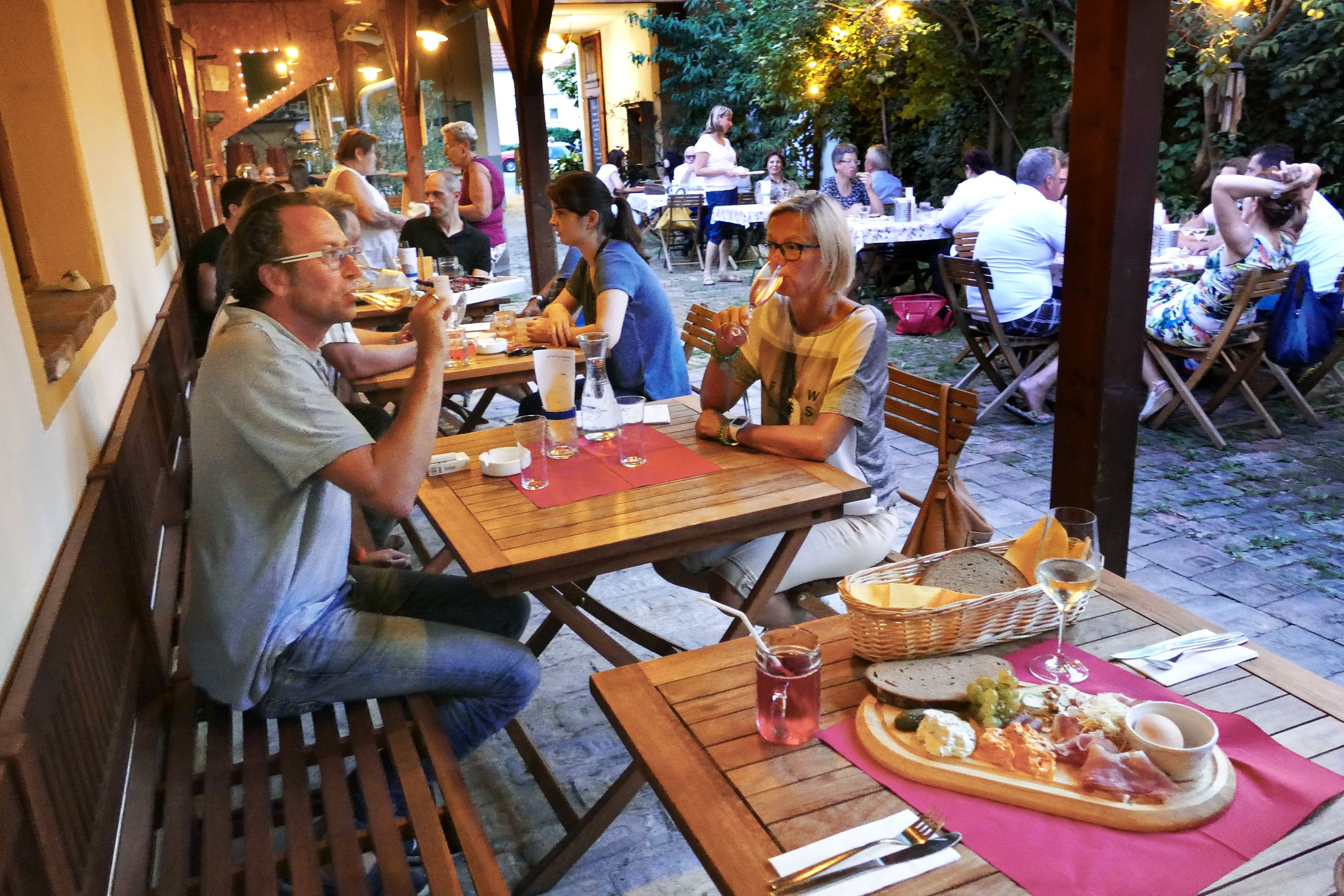 Az emberek fából készült asztaloknál ülnek egy hangulatos szabadtéri területen, és élvezik az ételeket és italokat.