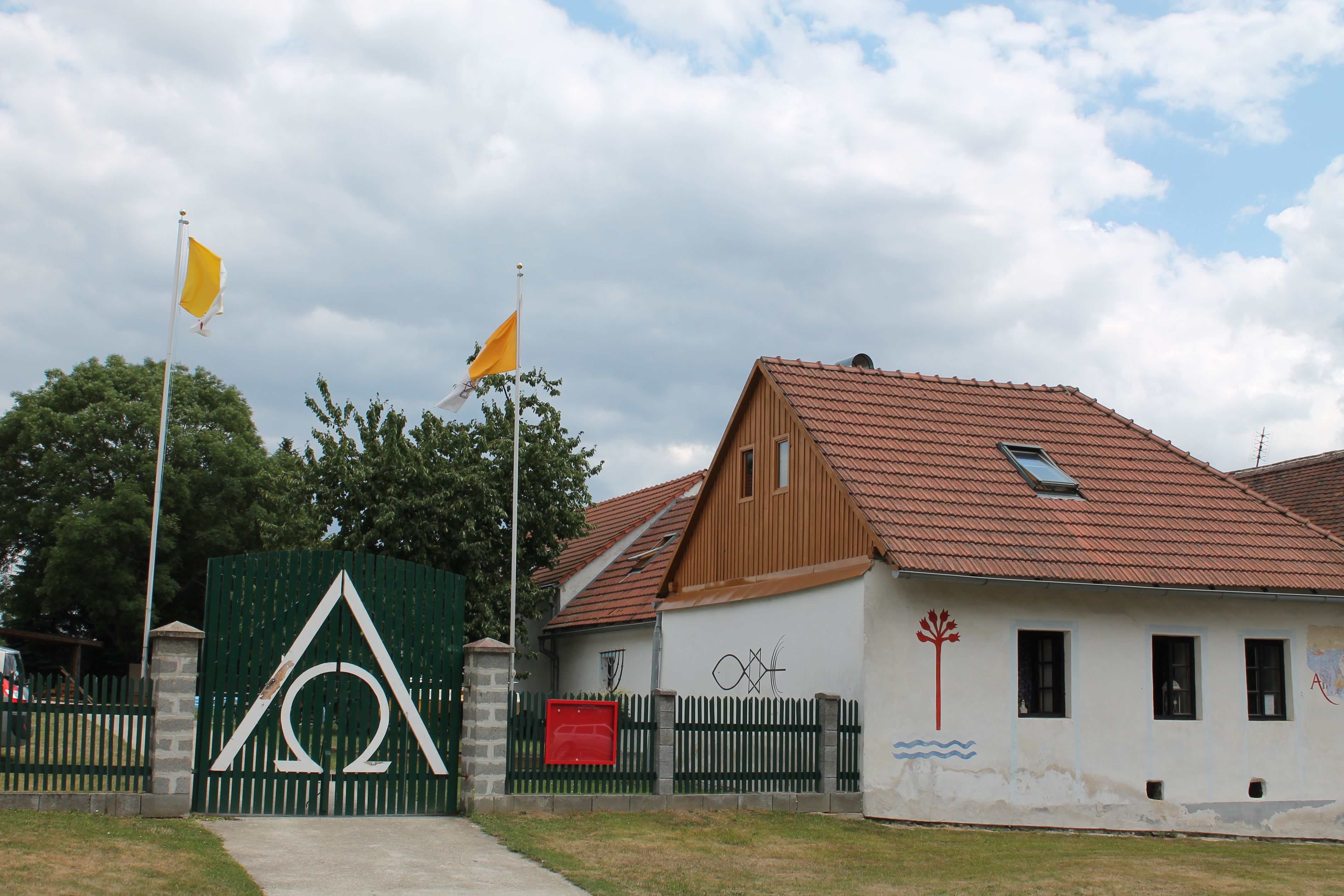 Egy ház piros tetőcserepekkel, zöld kerítés az Alpha Omega szimbólummal, két sárga zászló és falfestmények.