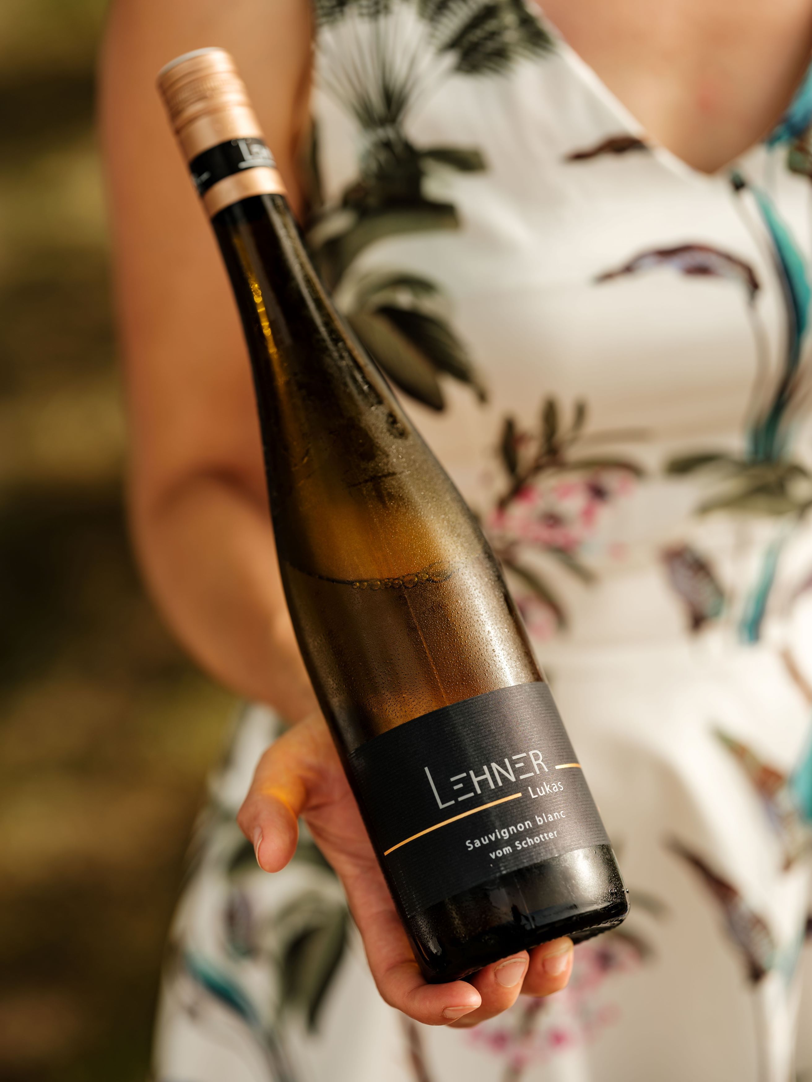 Az egyik személy egy üveg bort tart a kezében, amelyen a "Lehner Lukas Sauvignon blanc vom Schotter" felirat szerepel.