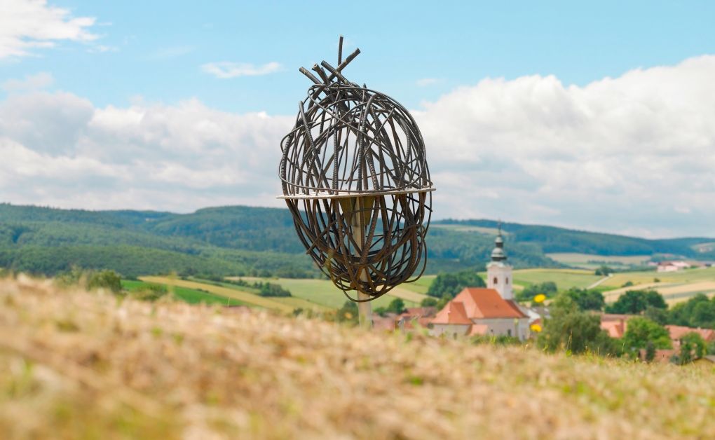 Fém művészeti installáció vidéki tájban, a háttérben templommal.