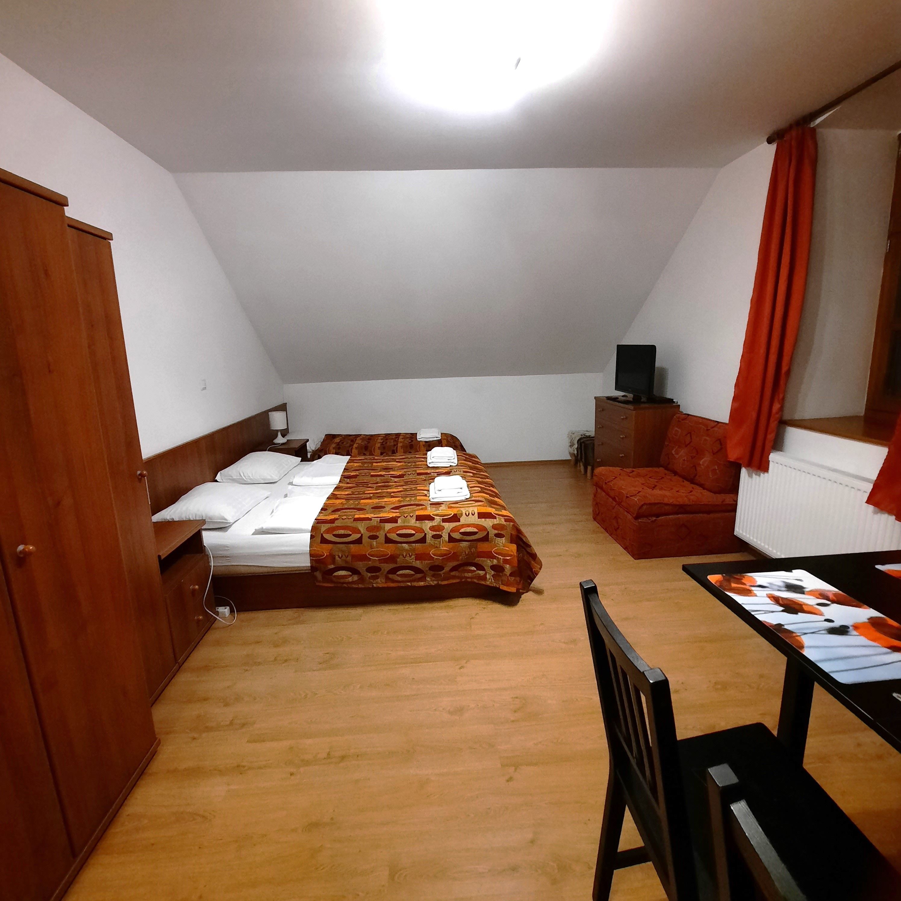 Egyszerűen berendezett szoba franciaággyal, kanapéval, asztallal és székkel. A szobát fából készült bútorok és narancssárga színű függönyök uralják.