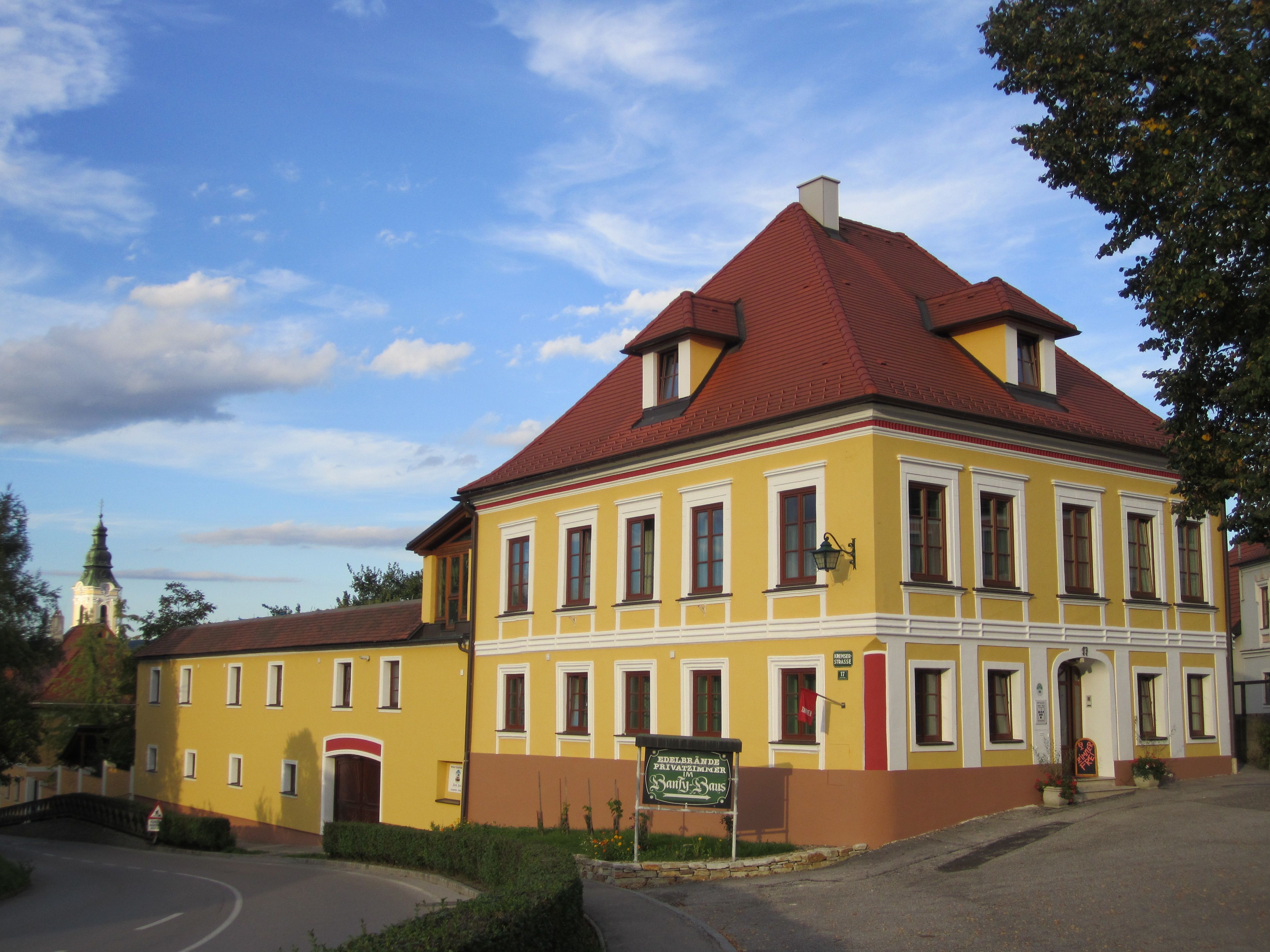 Sárga kétszintes épület piros tetővel és fehér ablakkeretekkel, mellette egy kisebb épület. A háttérben templomtorony.
