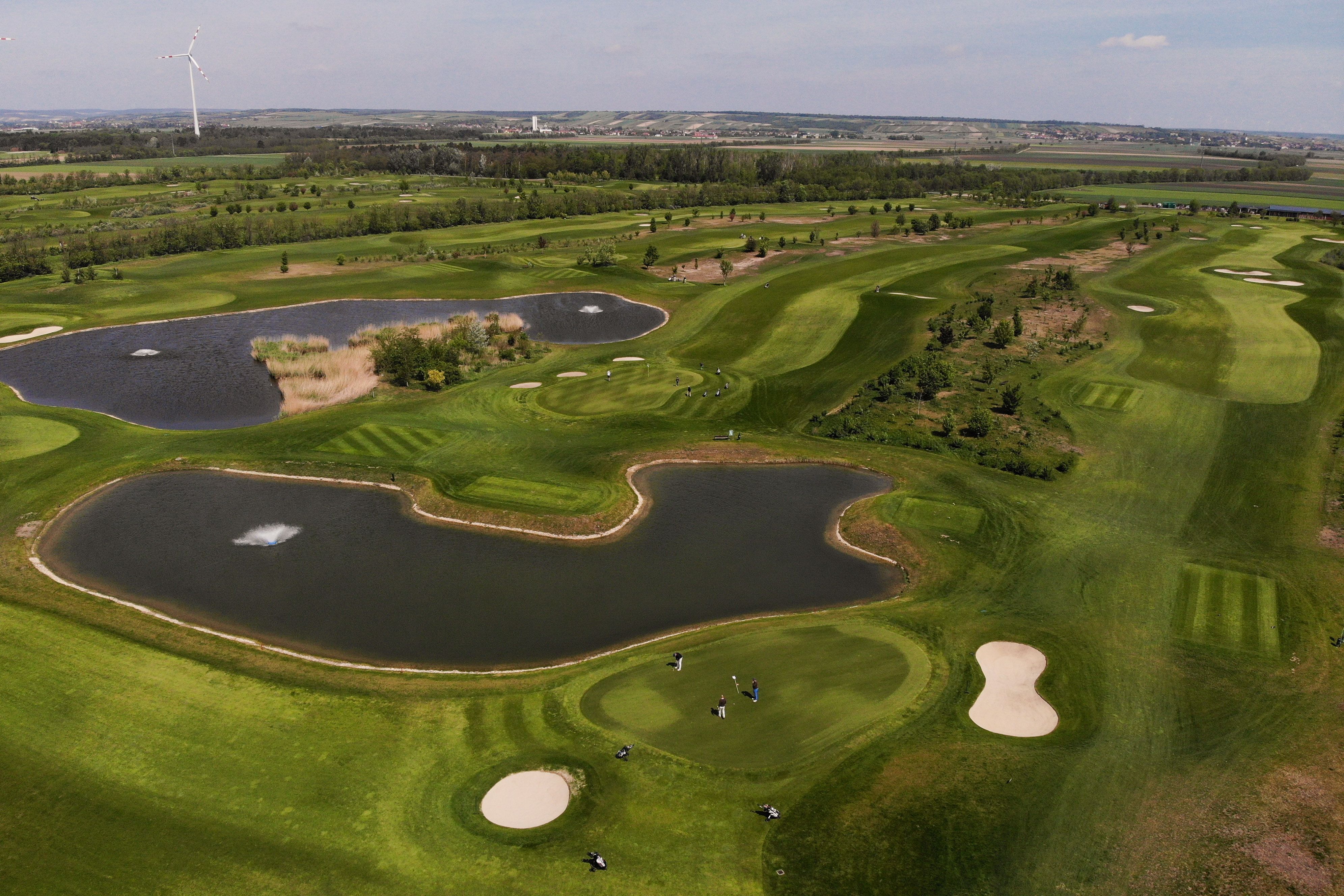 Légi felvétel egy golfpályáról, a háttérben tavakkal és szélturbinával.