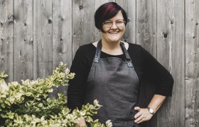 Egy szemüveges, kötényes nő áll mosolyogva egy bokor melletti fal előtt.