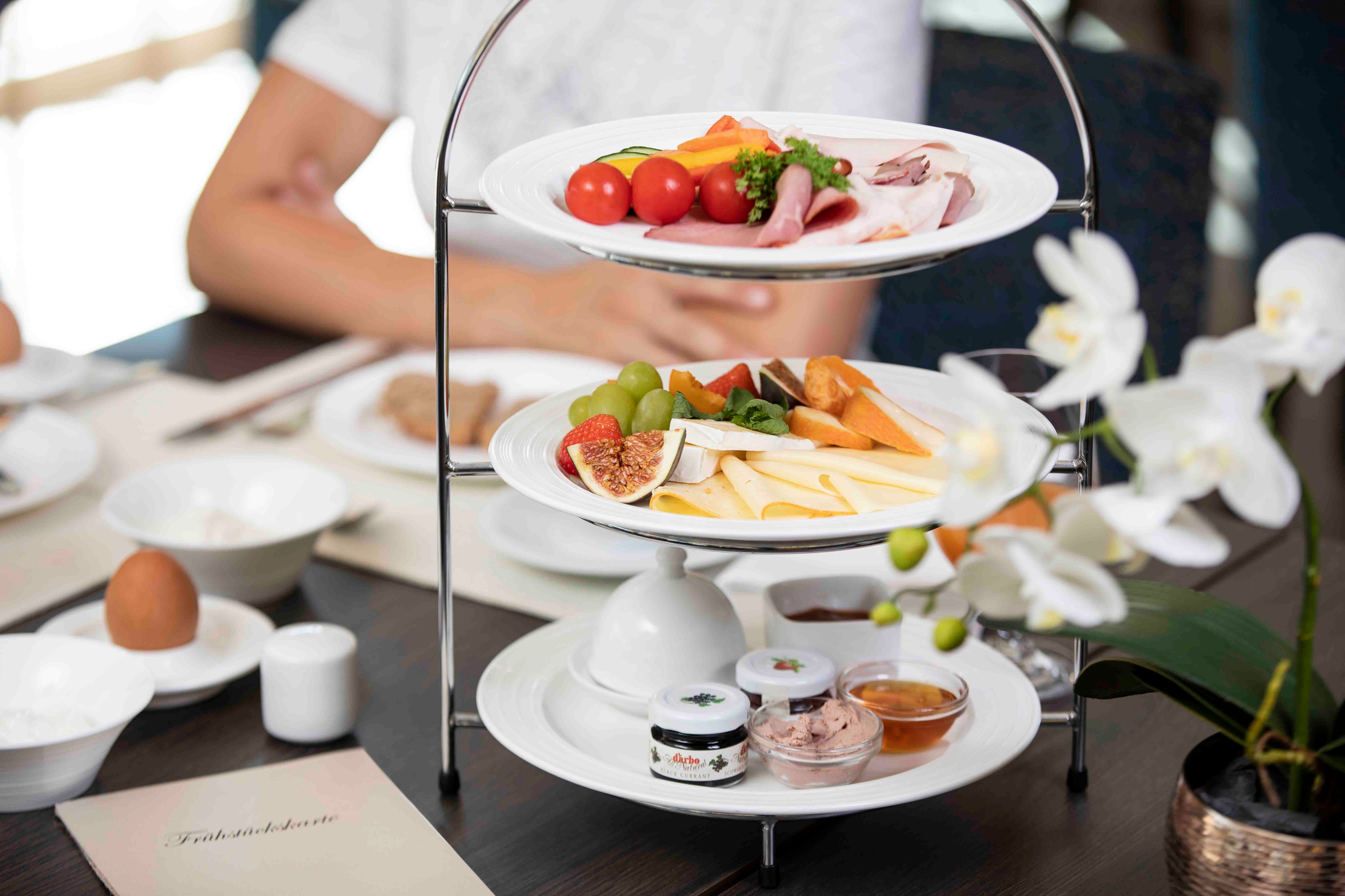 Egy reggelizőasztal, tele sajtokkal, felvágottakkal, gyümölcsökkel és kenőcsökkel. Az előtérben fehér orchidea.