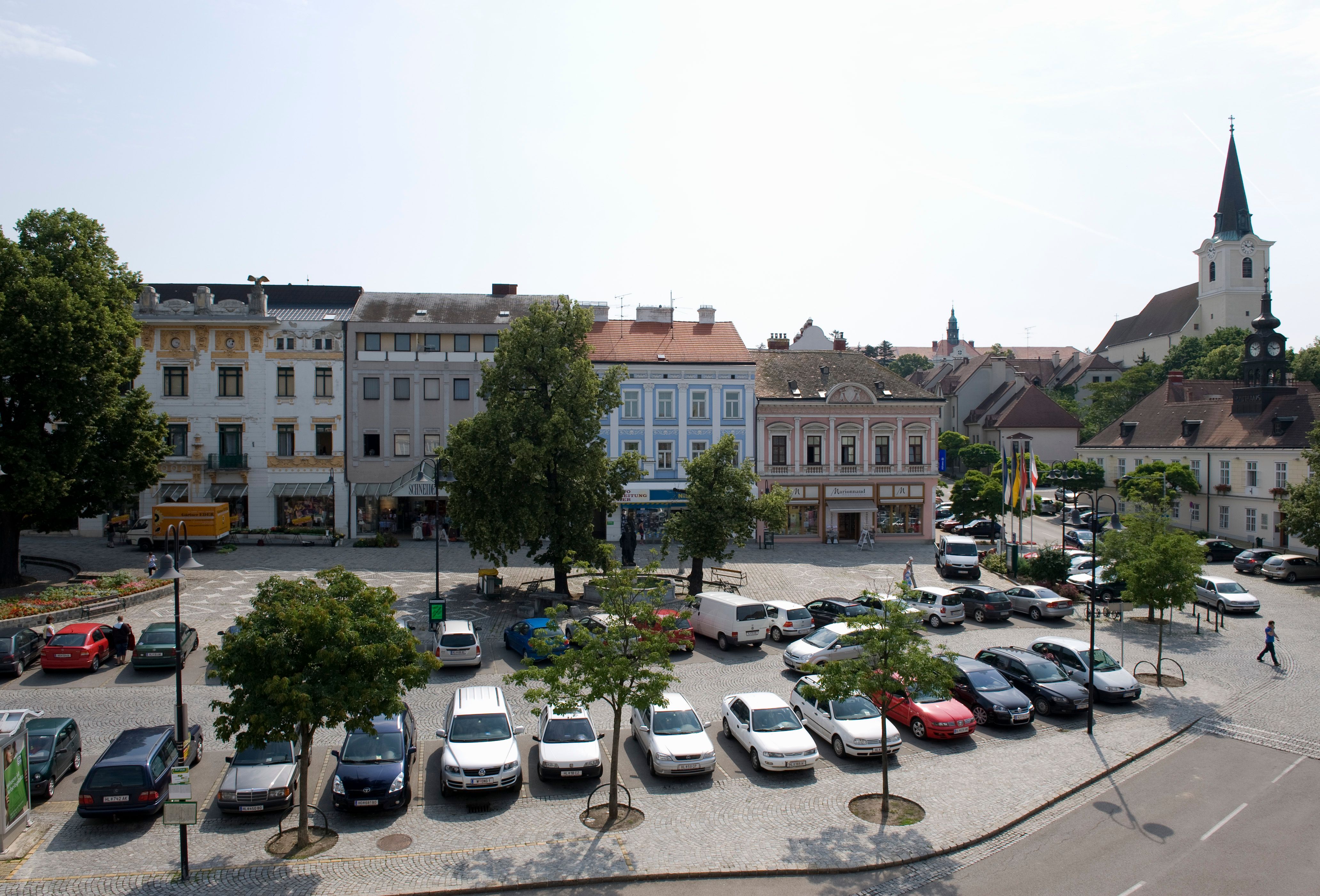 Hollabrunn város főtere parkoló autókkal, történelmi épületekkel és egy templommal a háttérben.