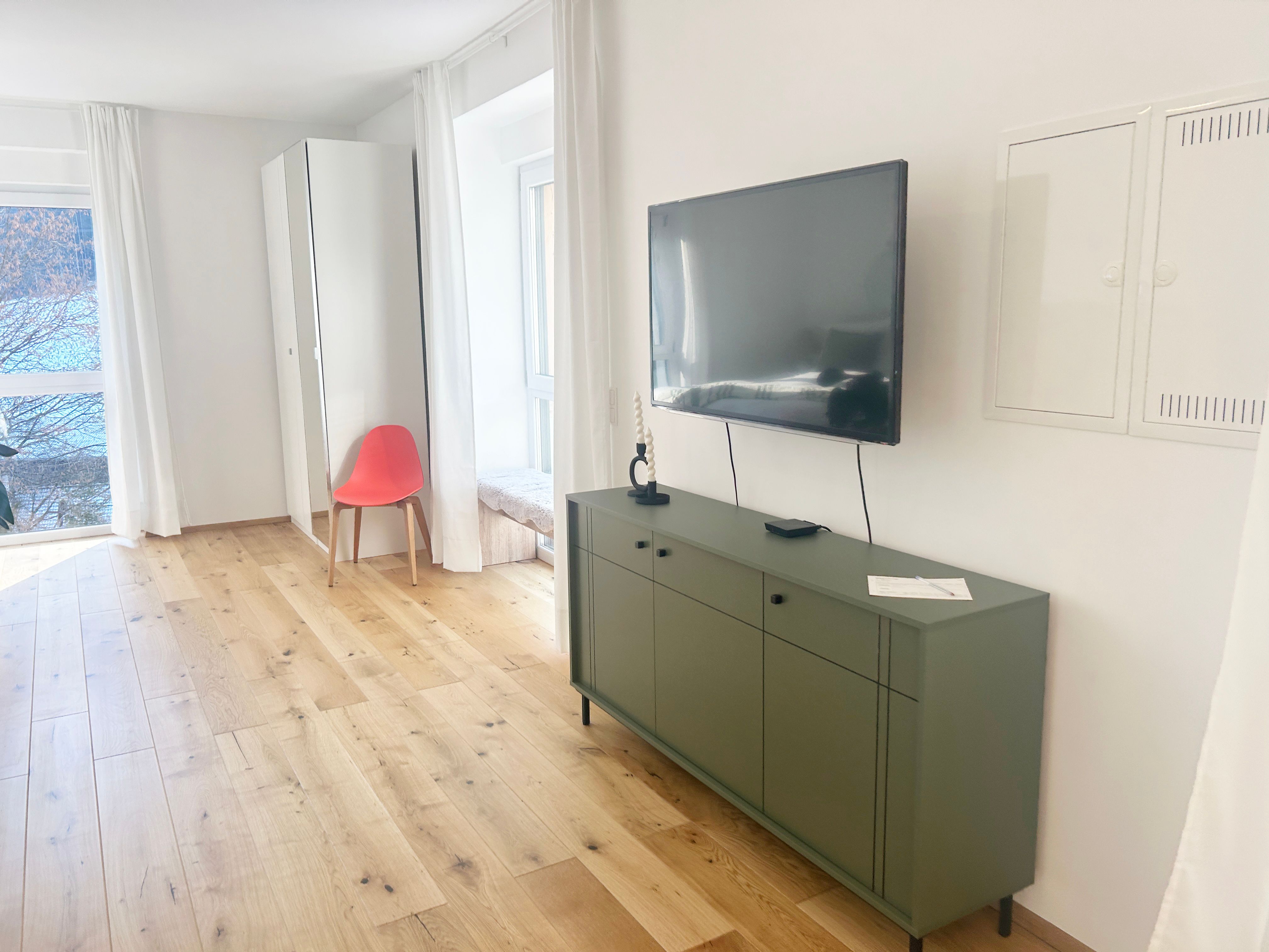 Modern nappali fapadlóval, zöld komóddal, falra szerelt televízióval és piros székkel a tükör előtt.