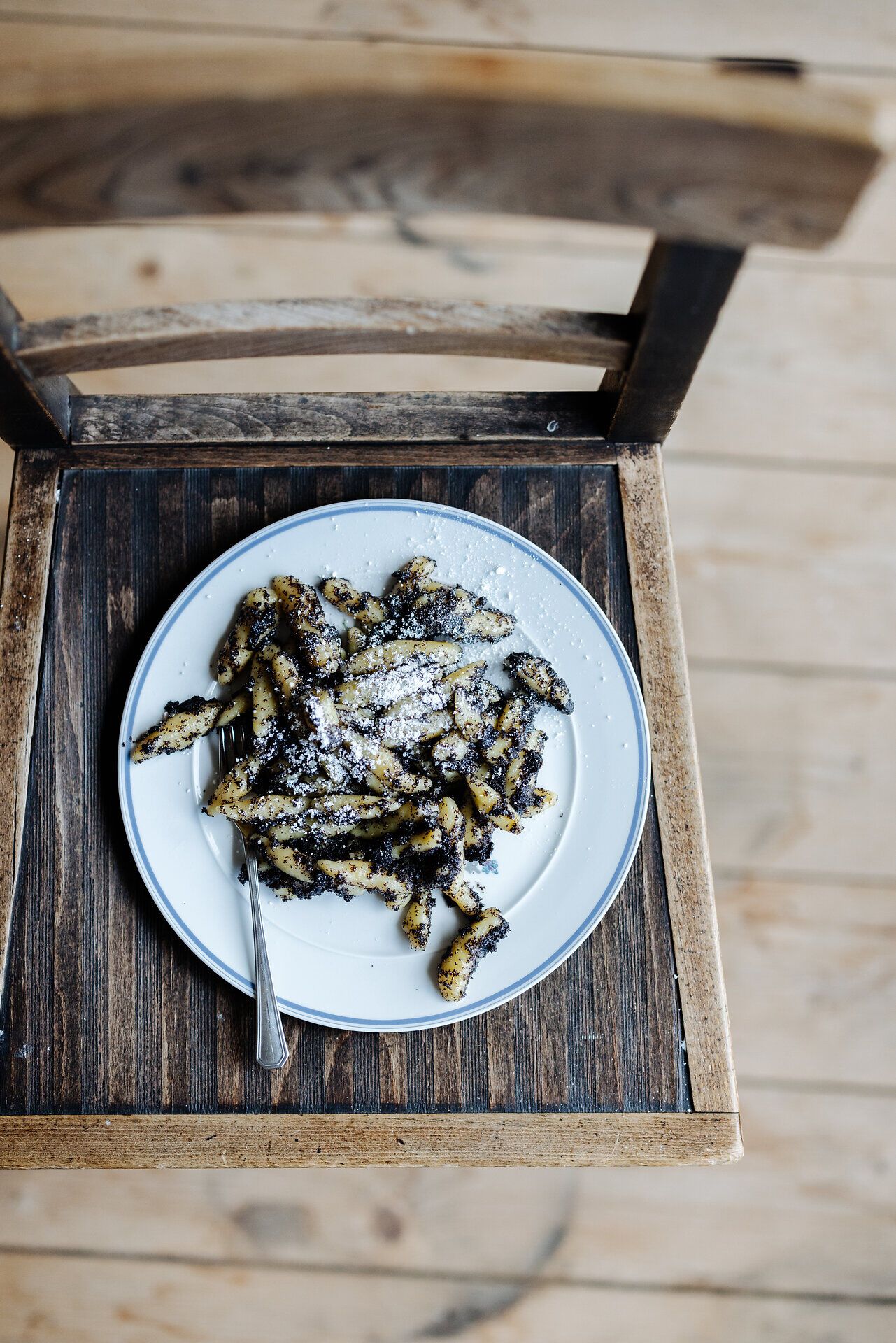 Ein köstlicher Teller Mohnnudeln, liebevoll angerichtet und mit Puderzucker bestäubt, lädt zum Genießen ein. Die rustikale Holzstuhlkonstruktion und der warme Holzboden schaffen eine einladende Atmosphäre, die zum Verweilen einlädt. Hier wird die Tradition der regionalen Küche lebendig.