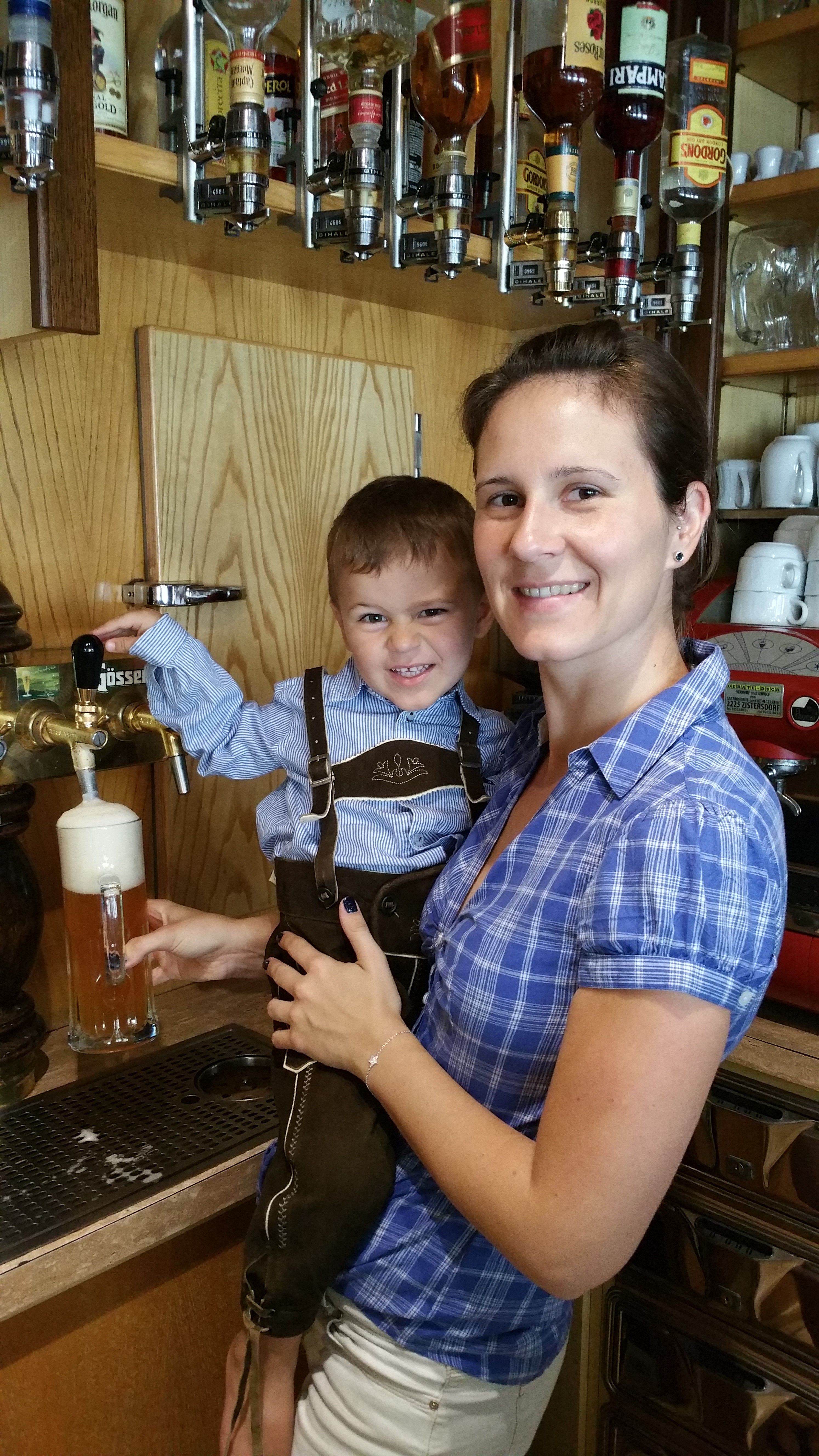 Egy nő egy sörcsapnál tart egy bőrnadrágos gyereket. A háttérben szeszesitalos palackok láthatók.