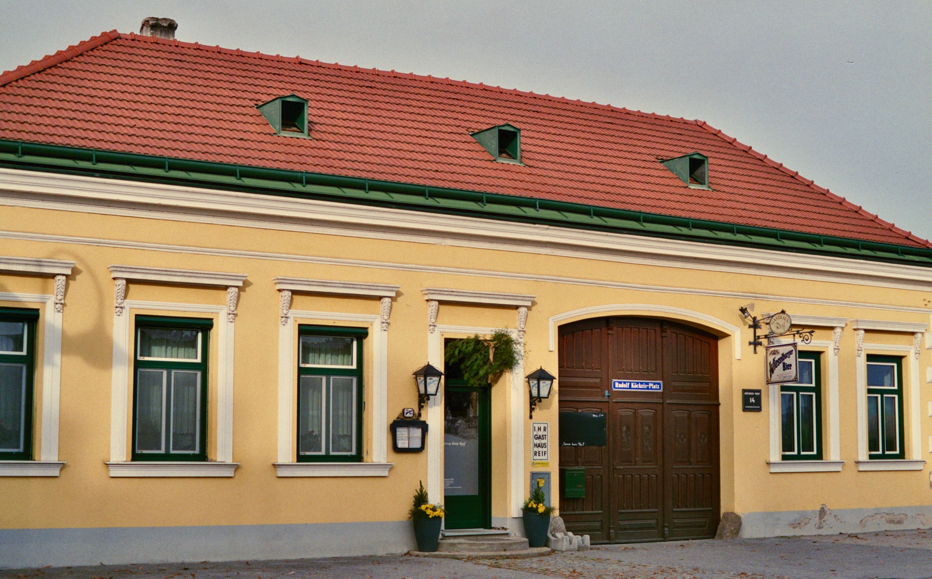 Egy sárga színű vendéglő külső nézete piros tetőcseréppel és zöld ablakkeretekkel.