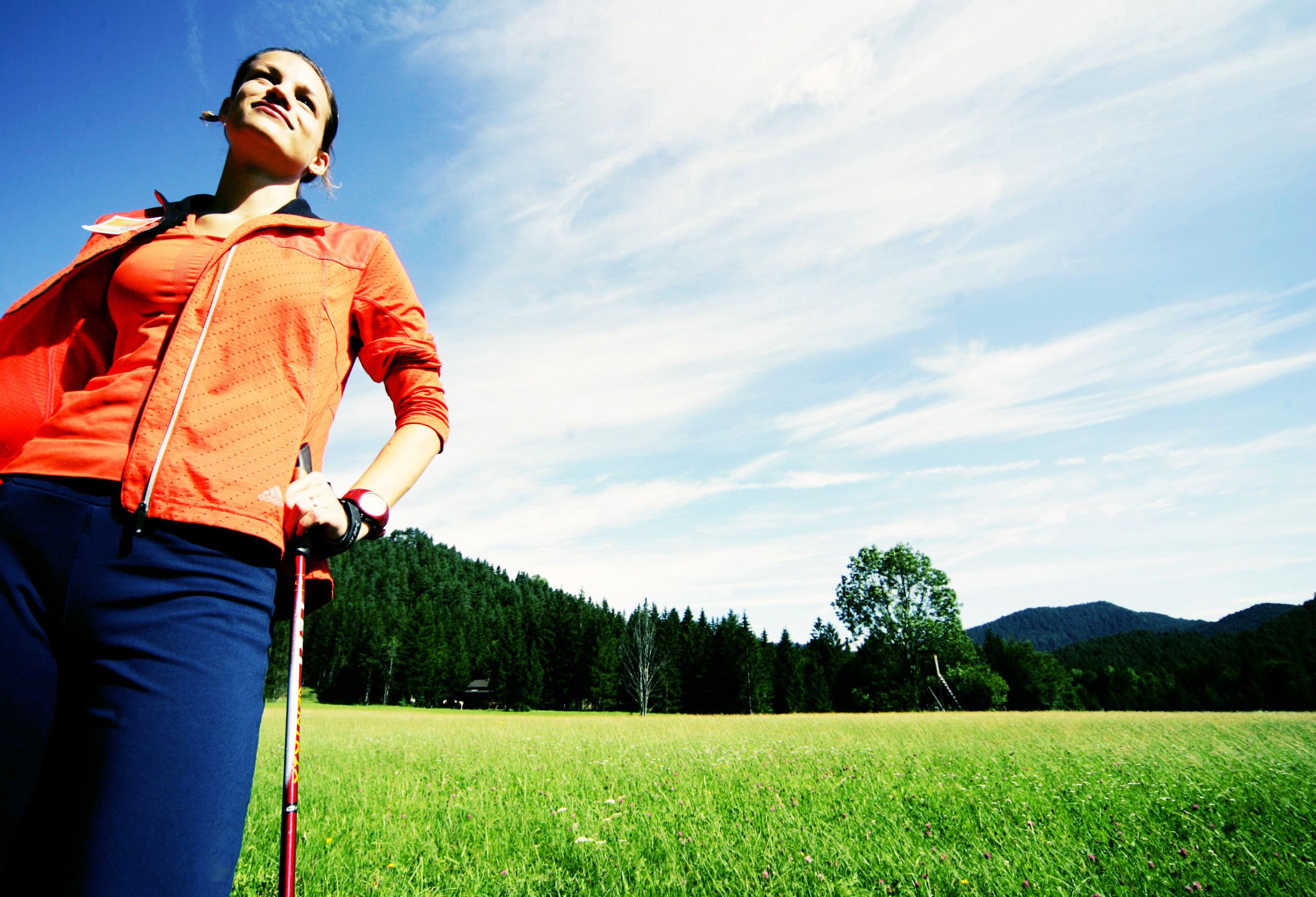 Nő sportruházatban, nordic walking botokkal egy erdő előtti réten.